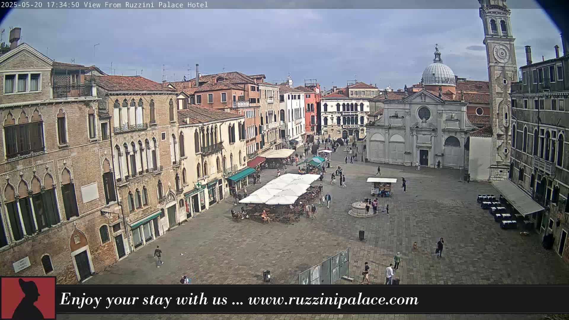 A large, paved square in Venice, Italy, is filled with people and outdoor seating under umbrellas, surrounded by old, multi-story buildings under a partly cloudy sky.
