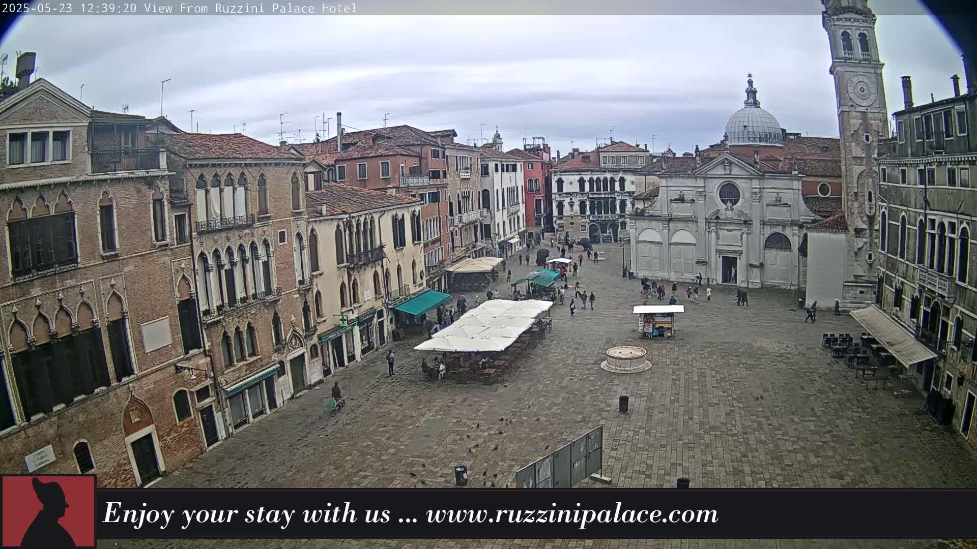 A mostly overcast day reveals a large, cobblestone town square in Venice, Italy, surrounded by historic buildings, with a few people and outdoor seating visible.