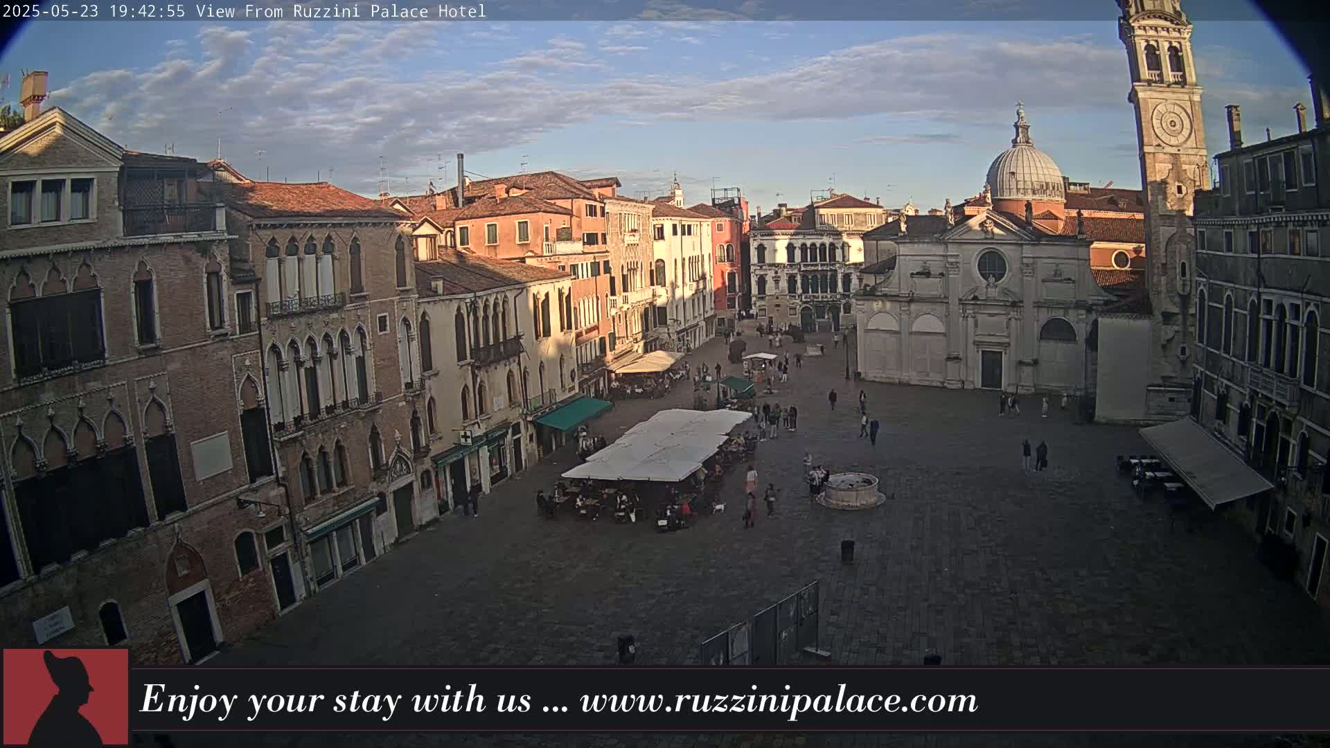 A mostly empty Venetian square is seen at dusk, surrounded by aged, multi-story buildings, one of which has a clock tower.