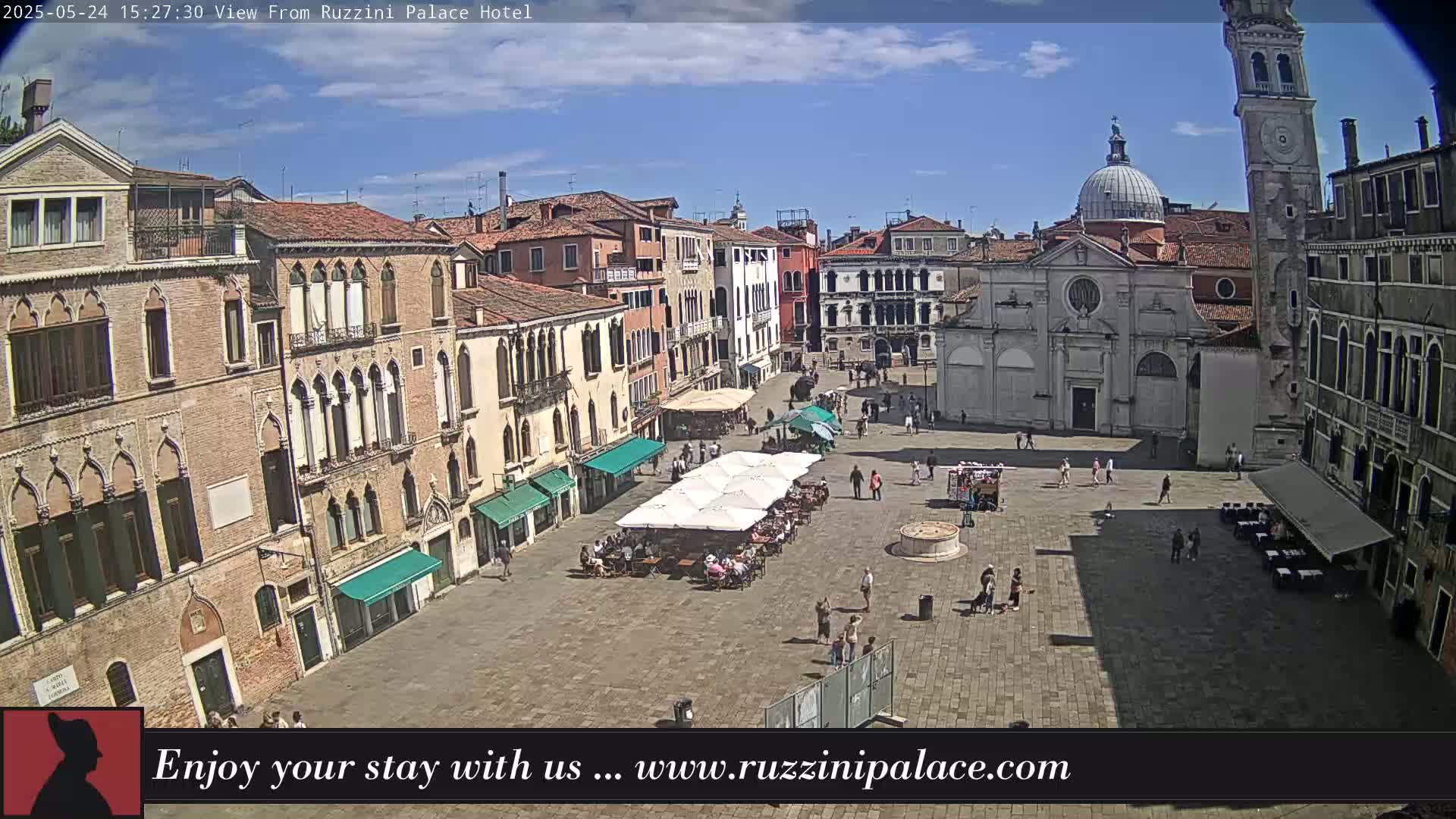 A sunny day reveals a large, paved square in Venice, Italy, surrounded by historic buildings, with several people walking and some seated at outdoor cafes under umbrellas.
