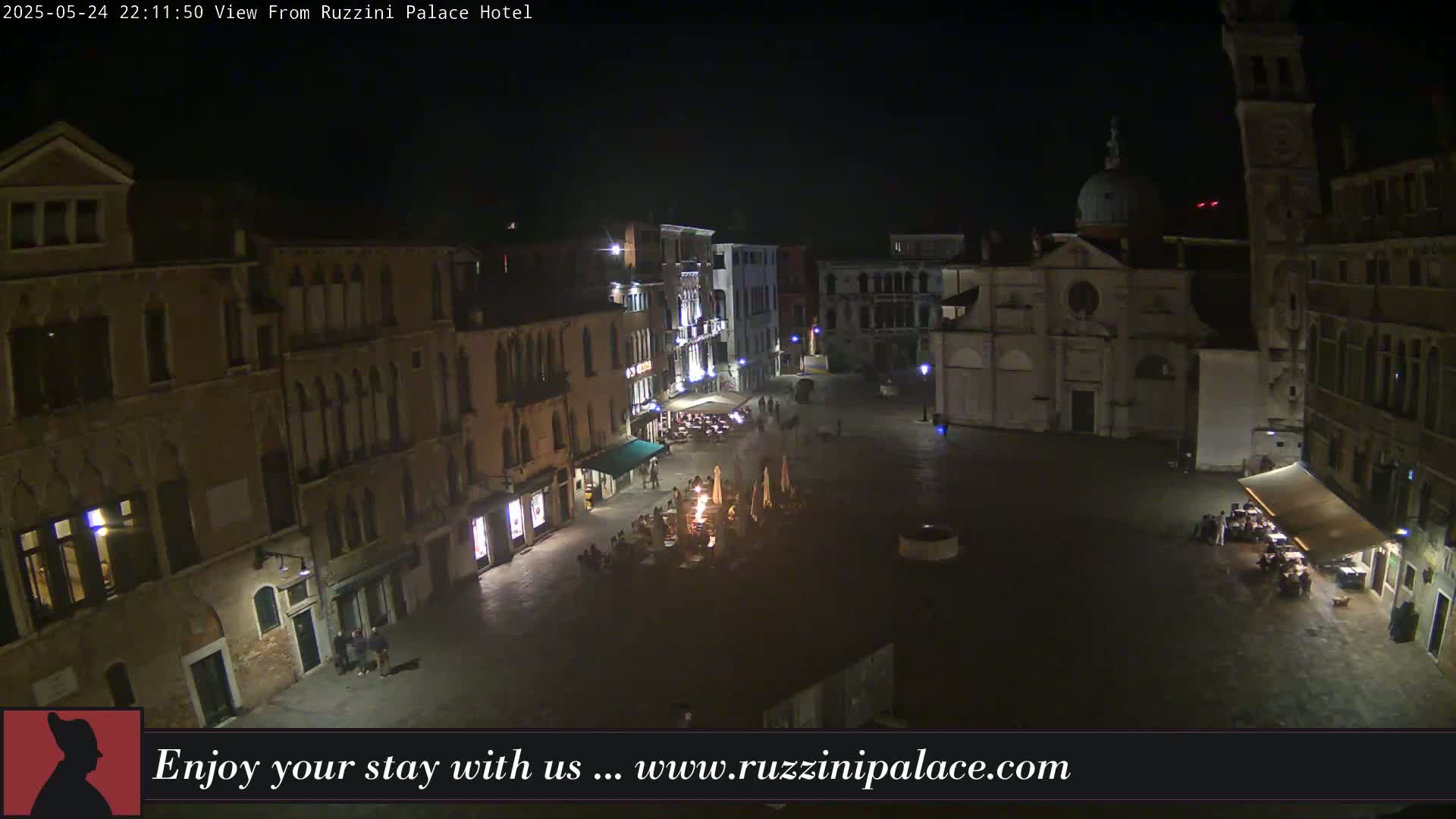 A nighttime aerial view shows a dimly lit square in Venice, surrounded by old buildings, with several outdoor dining areas occupied by people.