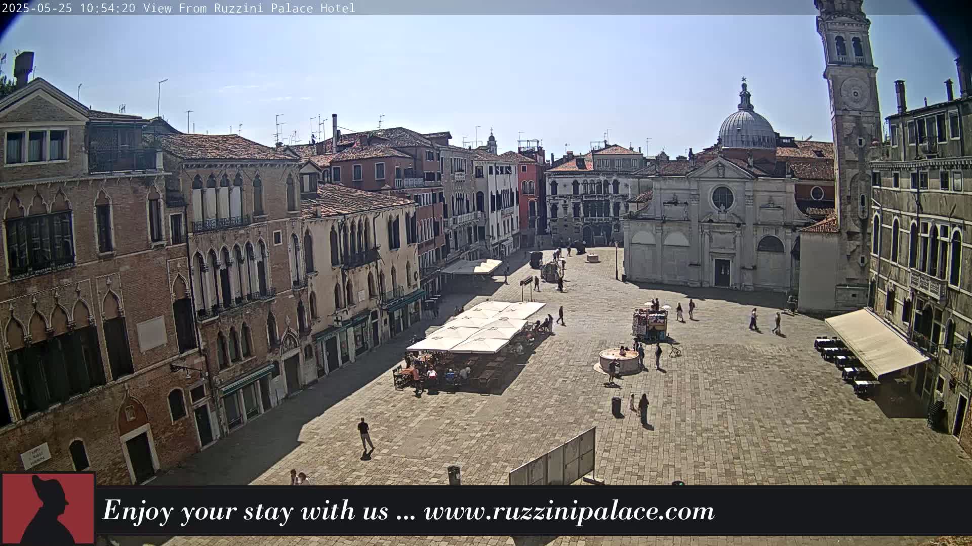A sunny Venetian plaza, surrounded by historic buildings, features a few people and outdoor seating under umbrellas.