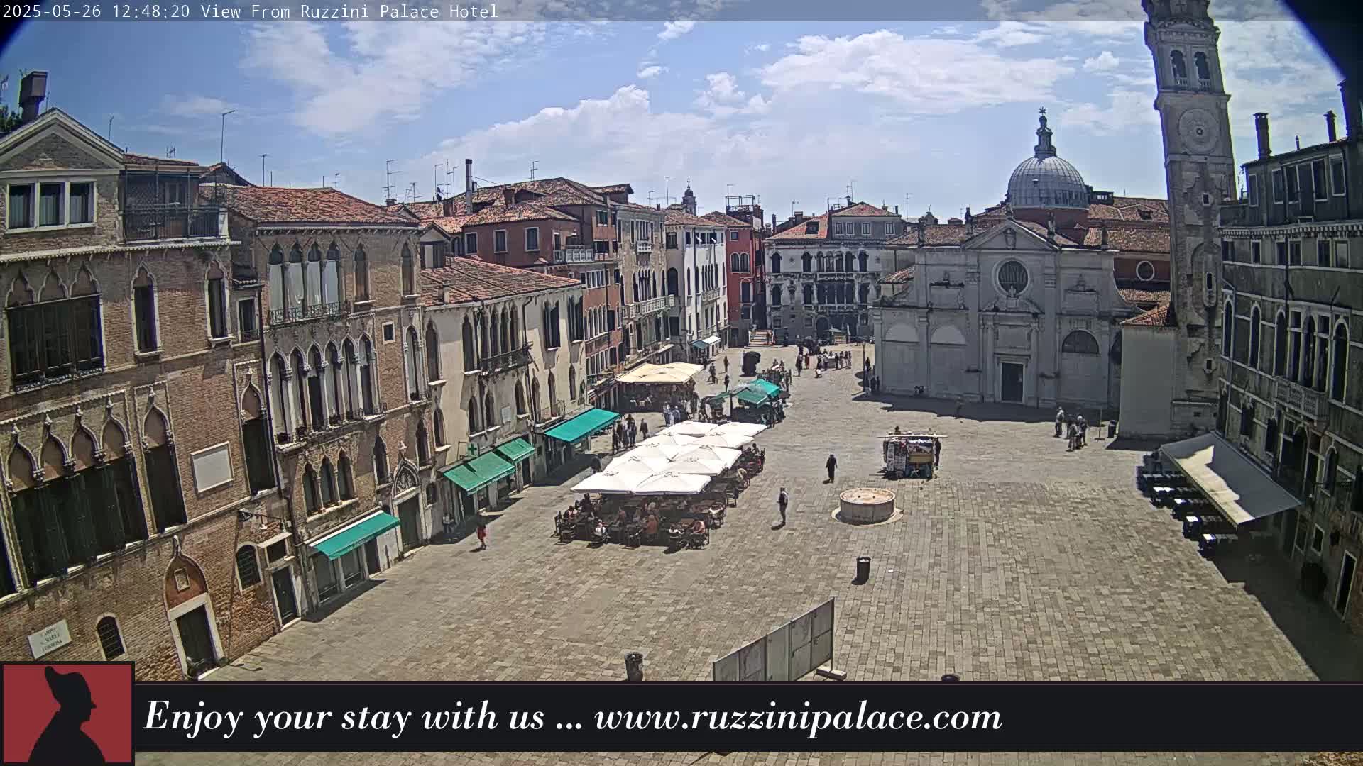 A sunny, partly cloudy day overlooks a large, paved square in Venice, Italy, surrounded by historical buildings, with several outdoor café umbrellas and a few people scattered throughout.