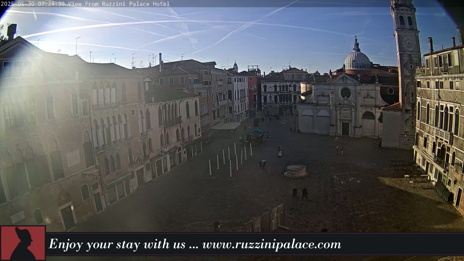 A sunny, clear morning view overlooking a large, mostly empty cobblestone square in Venice, surrounded by historic buildings, one of which has a domed roof and a tall clock tower.