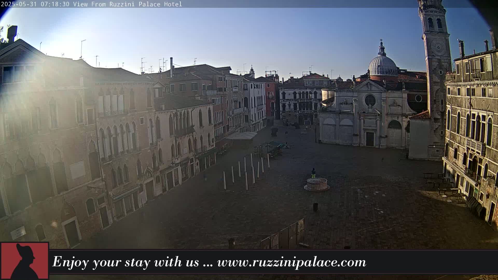 A mostly empty cobblestone plaza is surrounded by aged buildings under a clear, bright sky.