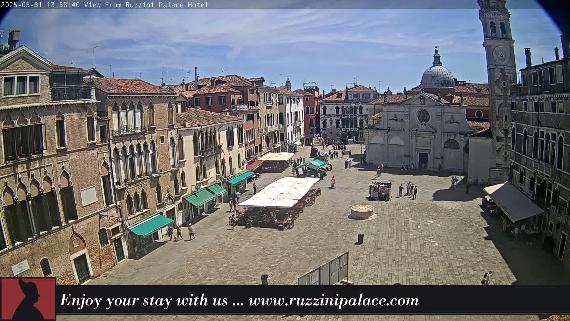 A sunny day in a large, paved European town square surrounded by historic buildings, with several people and outdoor seating areas visible.