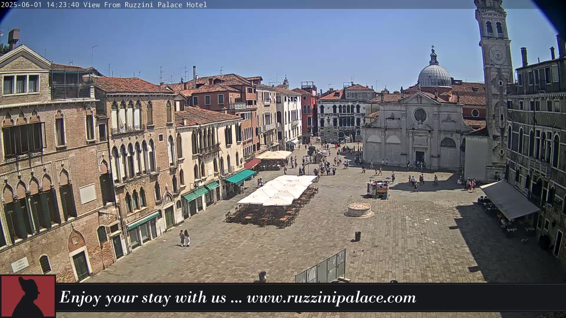 A sunny day in a large, cobblestone plaza in Venice, Italy, surrounded by old buildings, with several people and outdoor seating visible.