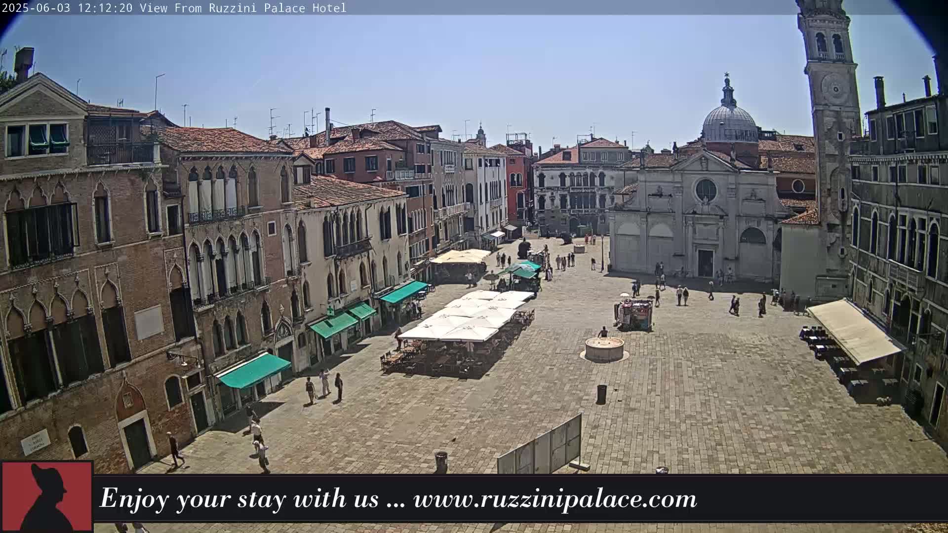 A sunny day in a large, paved European town square, surrounded by historic buildings, with several people walking around and outdoor seating under umbrellas.