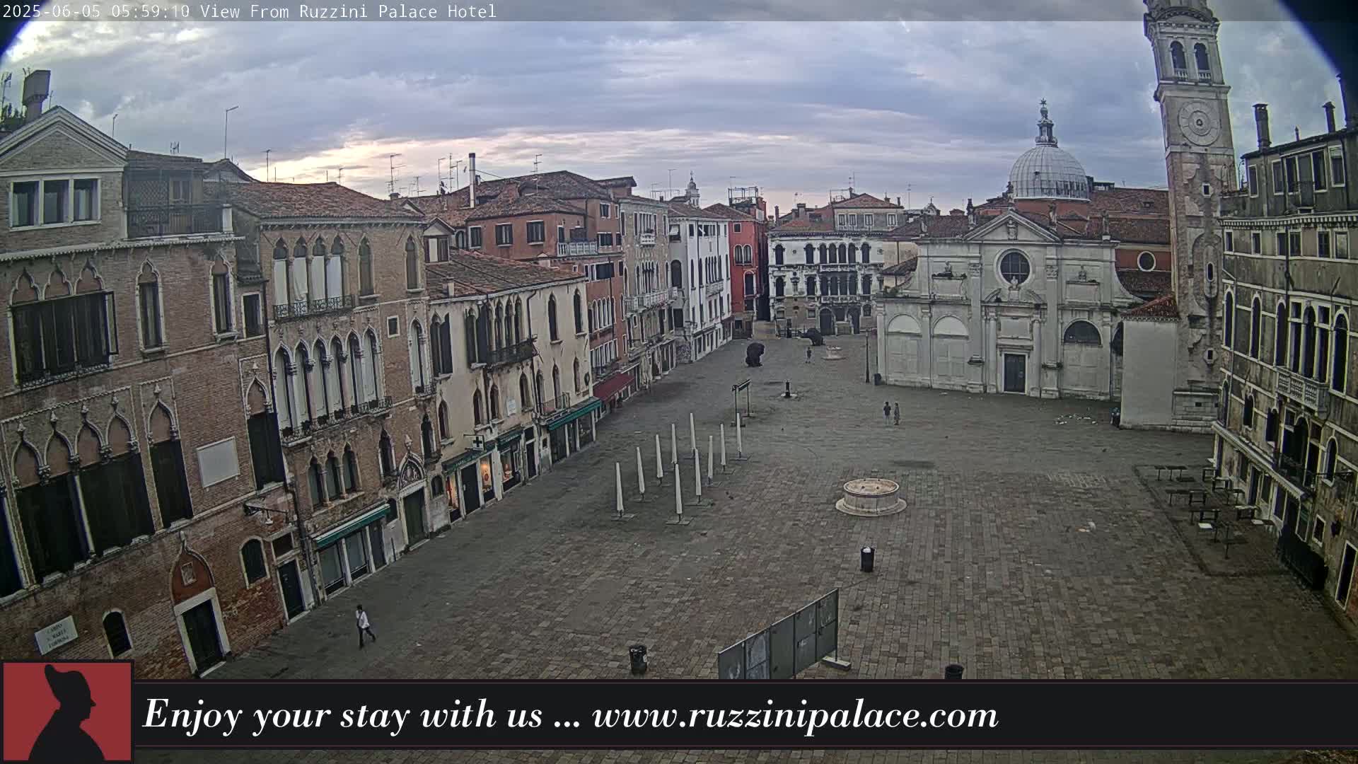 A mostly empty cobblestone square in Venice, under an overcast sky, is surrounded by historic brick buildings, one of which has a tall bell tower.