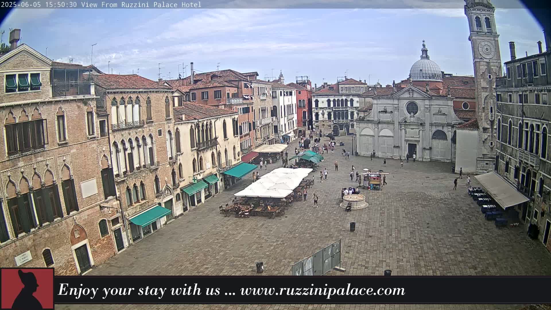 A partly cloudy day reveals a large, paved town square in Venice, Italy, surrounded by historic buildings, with several outdoor cafes set up with patrons.