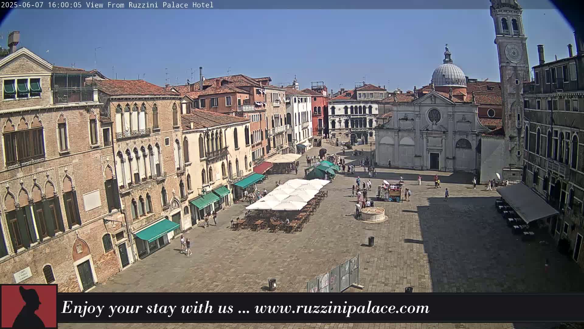 A sunny outdoor view of a large plaza in Venice, Italy, surrounded by historic buildings, with several people and outdoor seating areas visible.