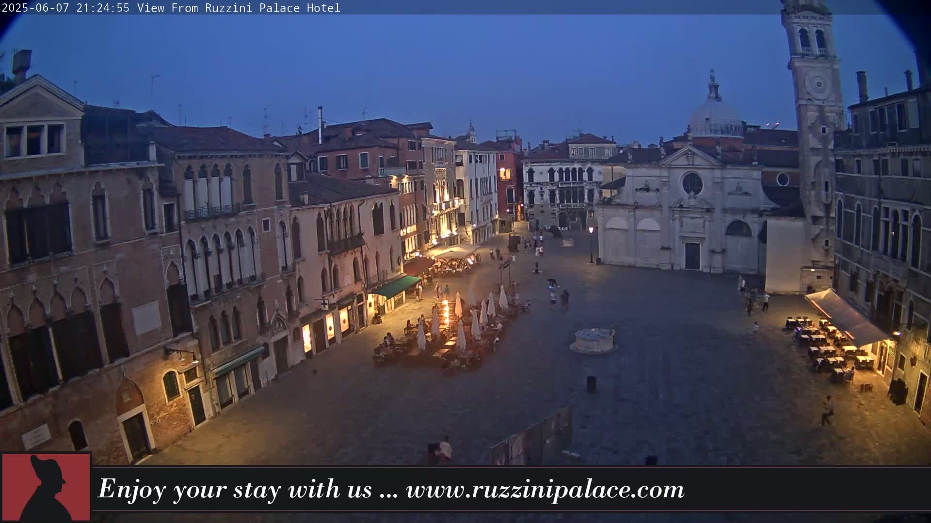 A twilight view of a large, mostly empty plaza in Venice, Italy, surrounded by old buildings, with several outdoor seating areas lit with warm light.