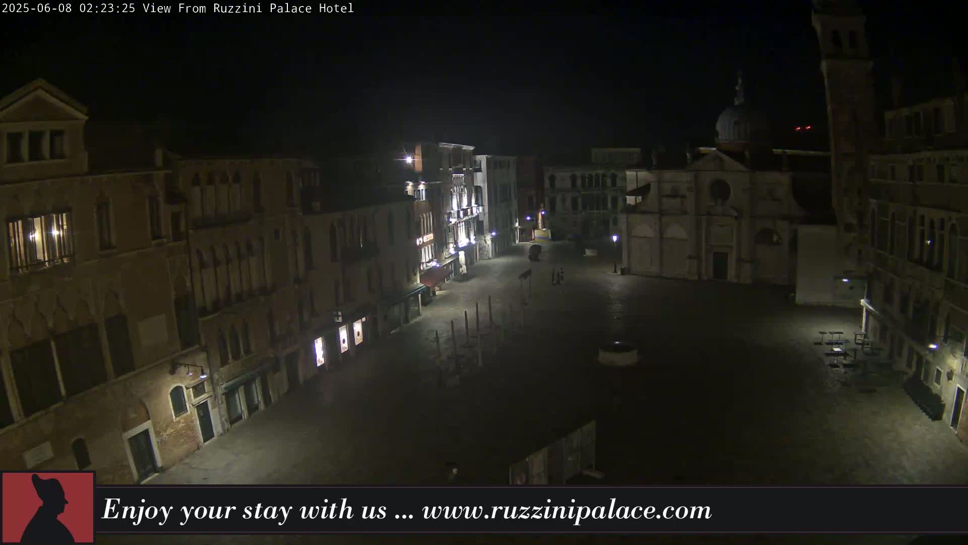A nighttime, mostly empty square in Venice, Italy, is surrounded by old buildings, with a few people and some outdoor furniture visible.
