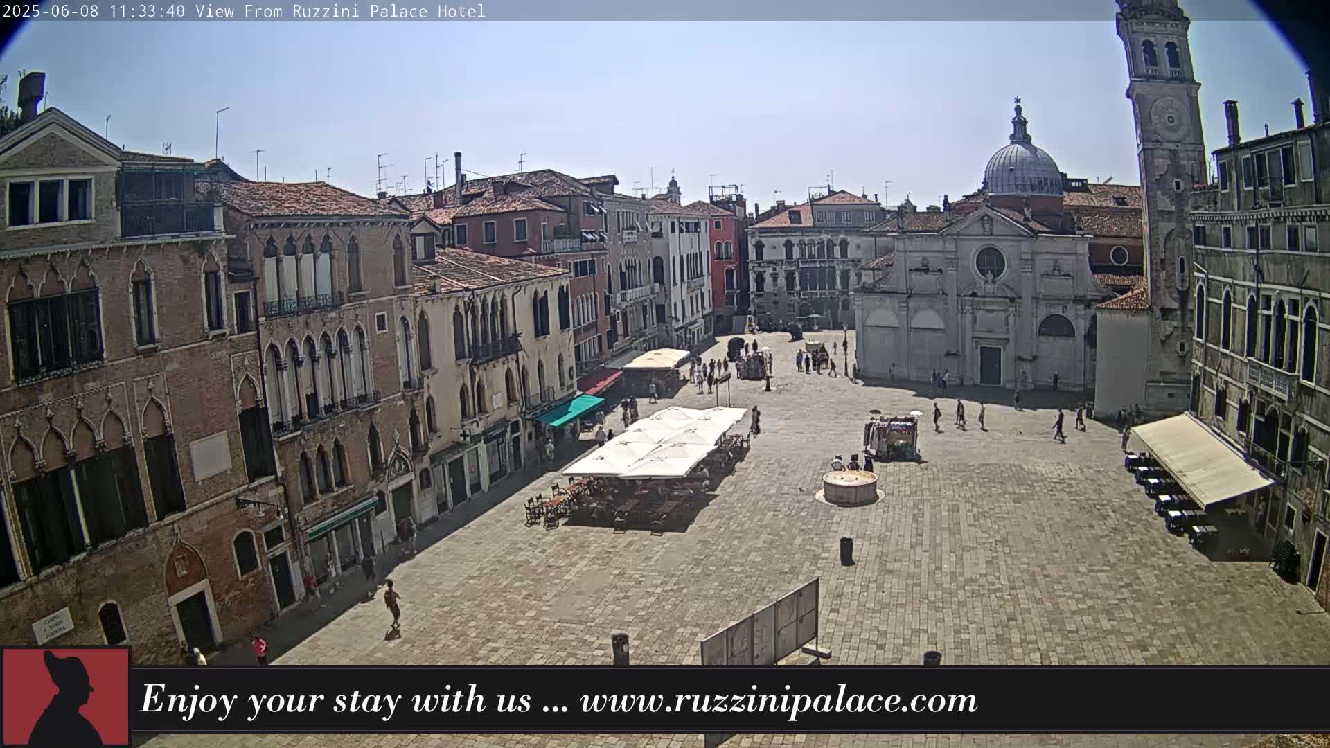 A sunny day in a large, paved square surrounded by historic buildings, with people scattered throughout and outdoor seating under umbrellas.