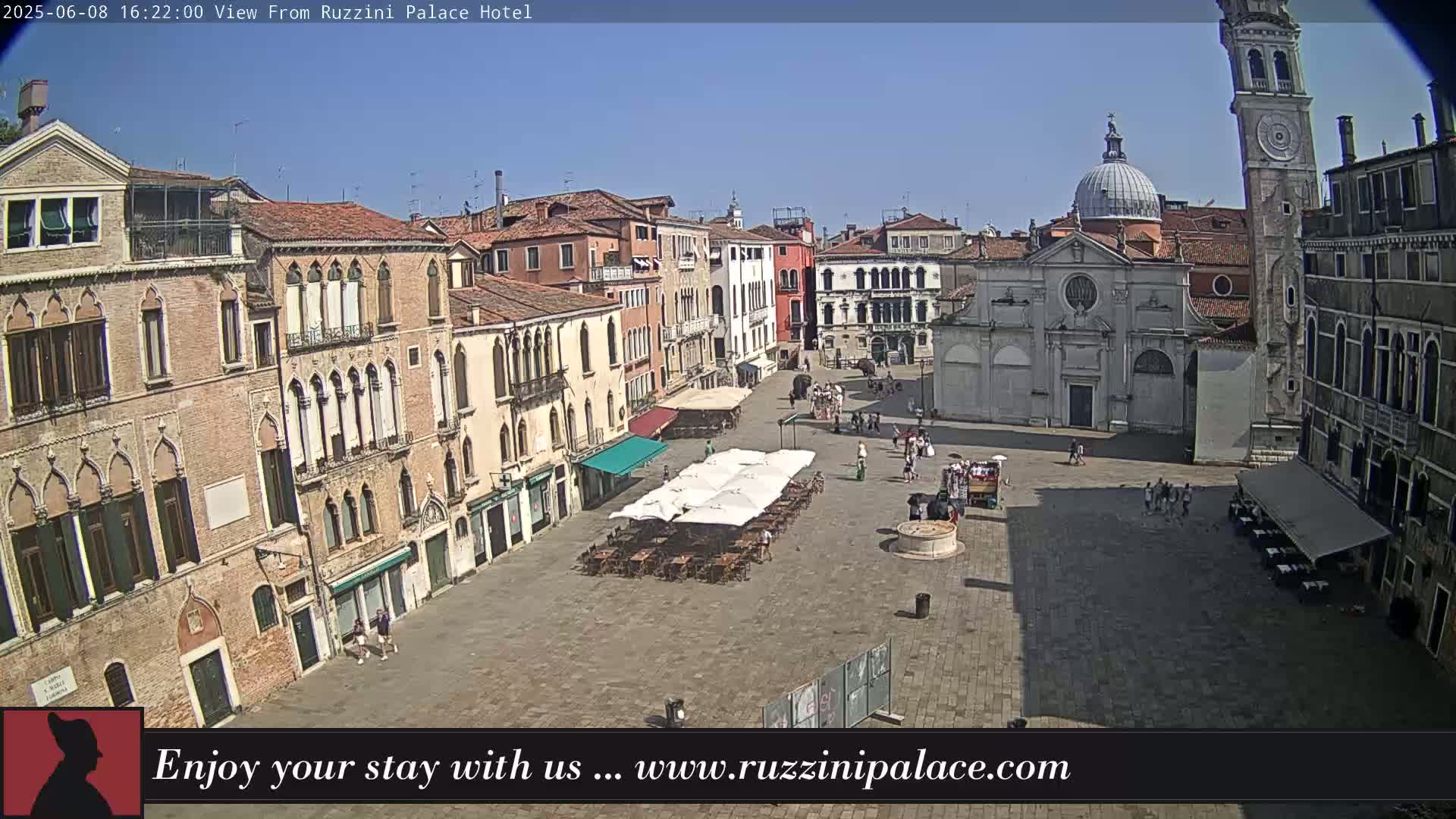 A sunny outdoor view of a large plaza in Venice, Italy, surrounded by old buildings, with several people and outdoor seating visible.