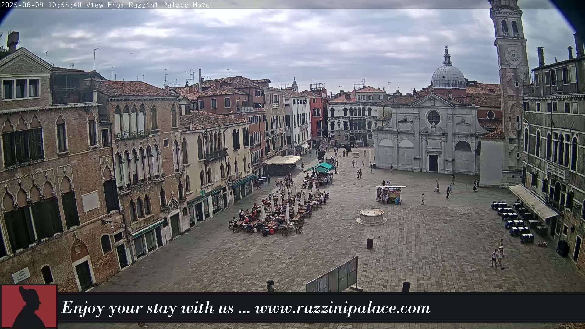 A mostly cloudy day shows an expansive cobblestone square in Venice, Italy, filled with outdoor seating and several buildings, including a church and a tall tower.