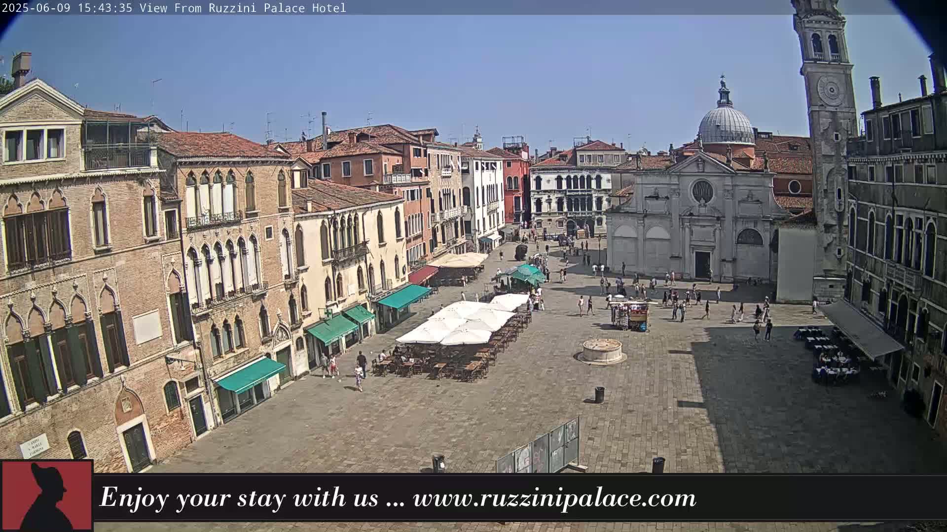 A sunny outdoor view shows a large town square in Venice, Italy, with several buildings surrounding it, some with outdoor seating, and people walking around.