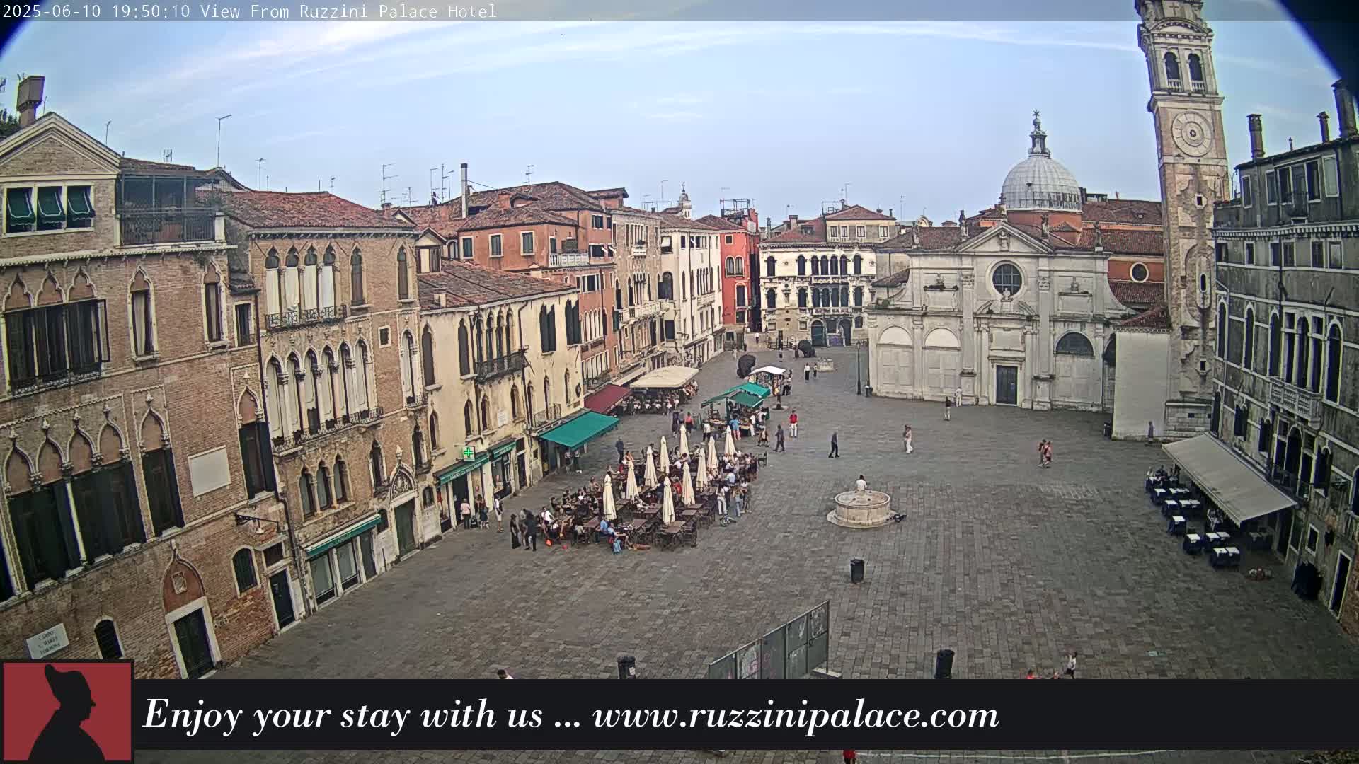 A mostly sunny outdoor view of a large, paved plaza in Venice, Italy, surrounded by historic buildings, with several people and outdoor seating areas visible.