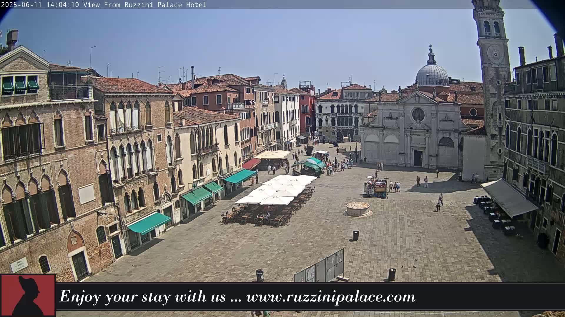 A sunny outdoor view shows a large, paved plaza surrounded by historic Venetian buildings, with some people and outdoor seating visible.