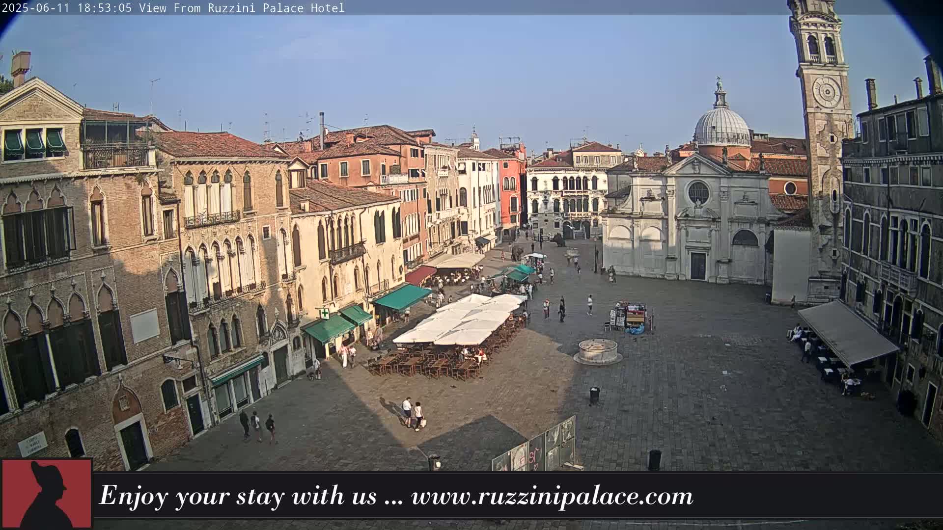 A sunlit Venetian square with several buildings, a church, and outdoor seating areas under umbrellas.