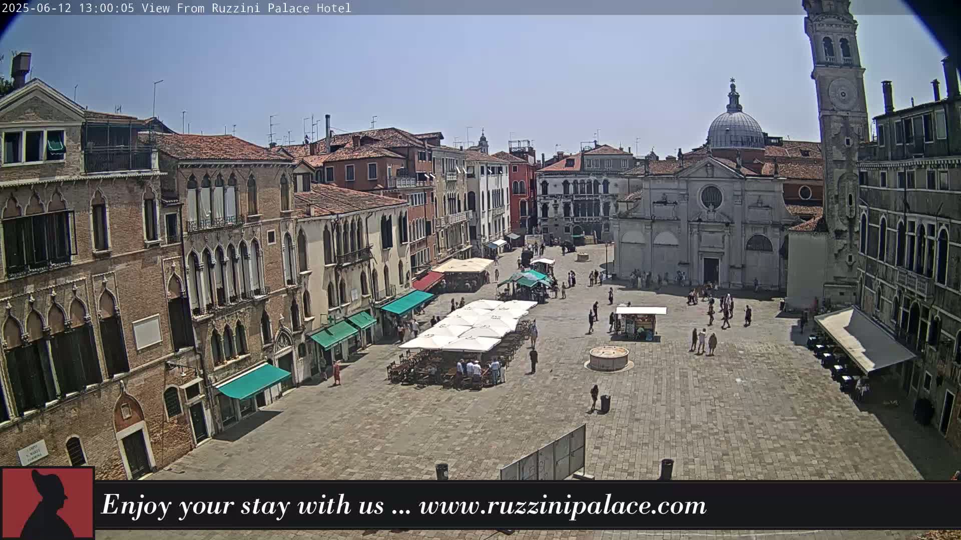 A sunny day reveals a large, paved square in Venice, Italy, surrounded by historic buildings, with several people and outdoor seating areas visible.