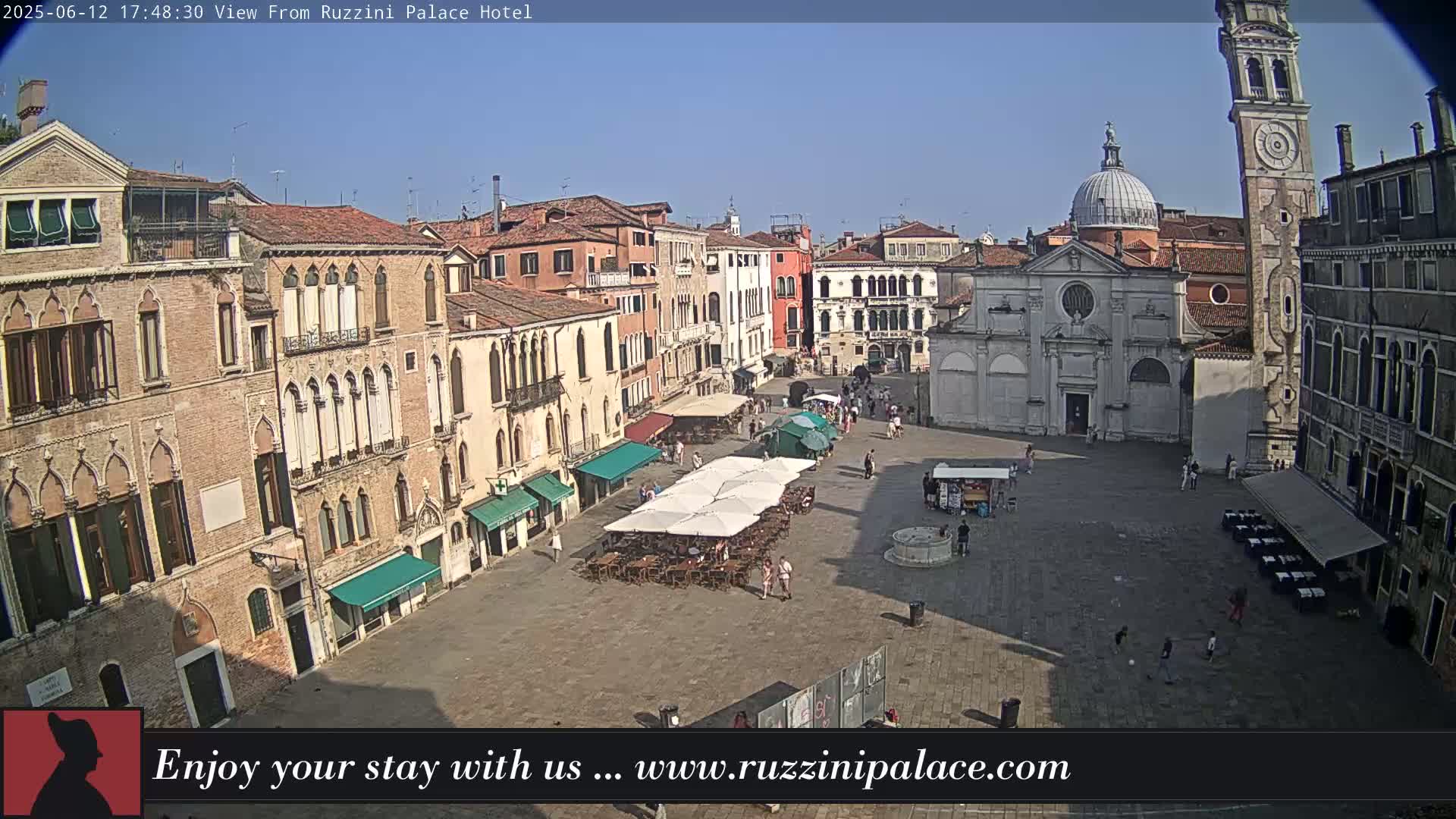 A sunny day reveals a large, paved European square surrounded by old buildings, with outdoor seating under umbrellas and a few people strolling.