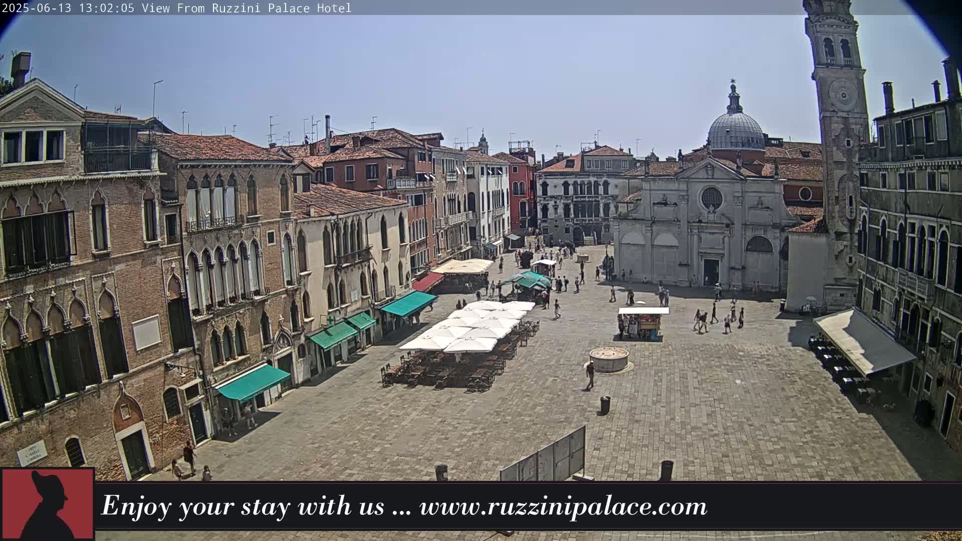 A sunny day in a large, paved European town square, surrounded by several-story buildings, with people walking around and some umbrellas set up for outdoor seating.
