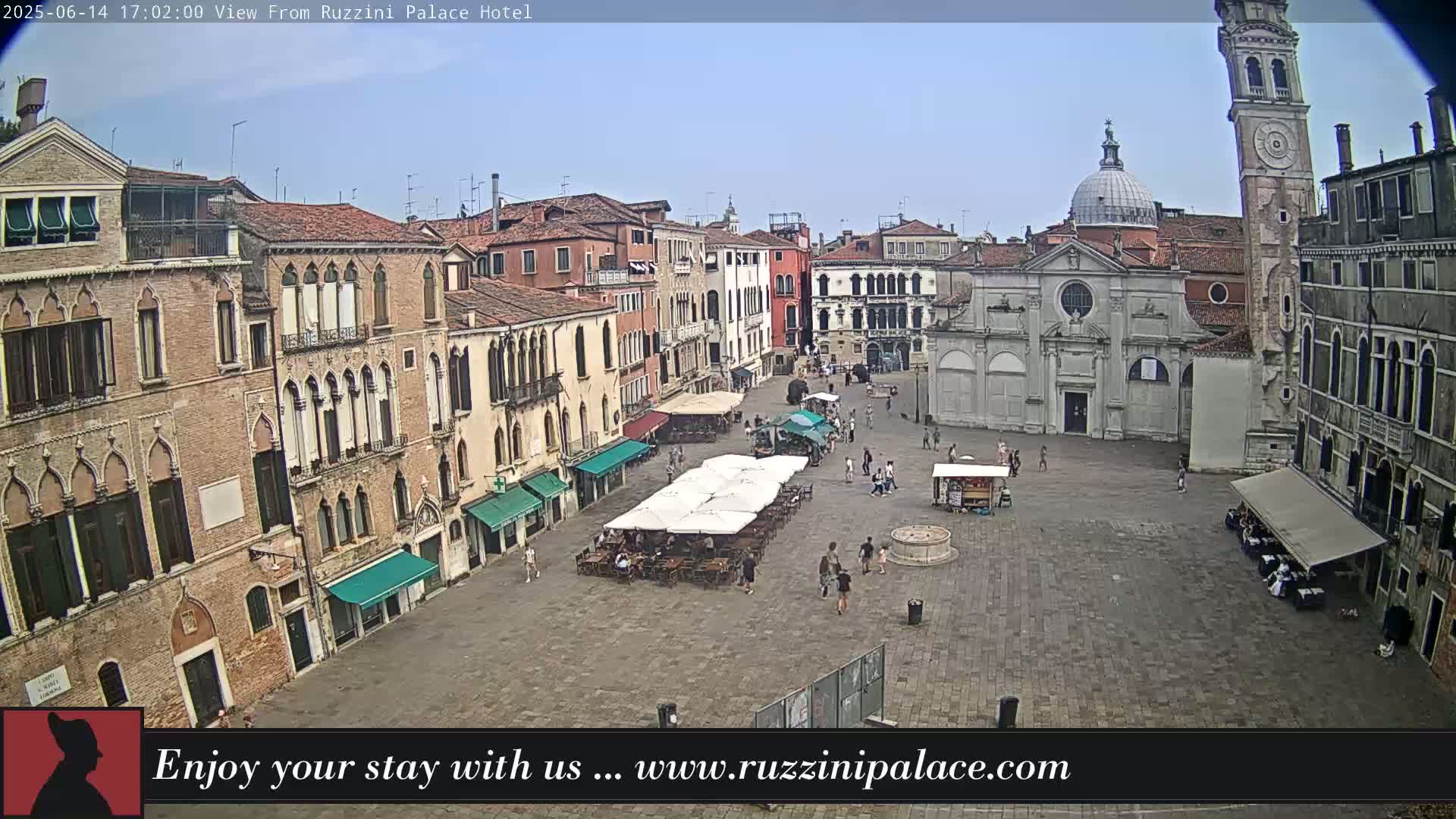 A sunny day reveals a large, paved square in Venice, Italy, surrounded by historic buildings, with outdoor seating areas and a few pedestrians visible.