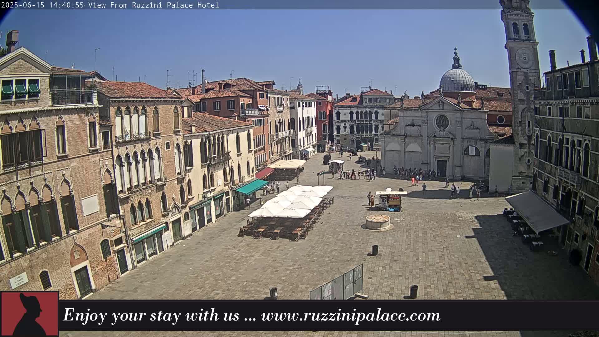 A sunny view of a large, paved square in Venice, Italy, surrounded by historic buildings, with several people and outdoor seating areas.