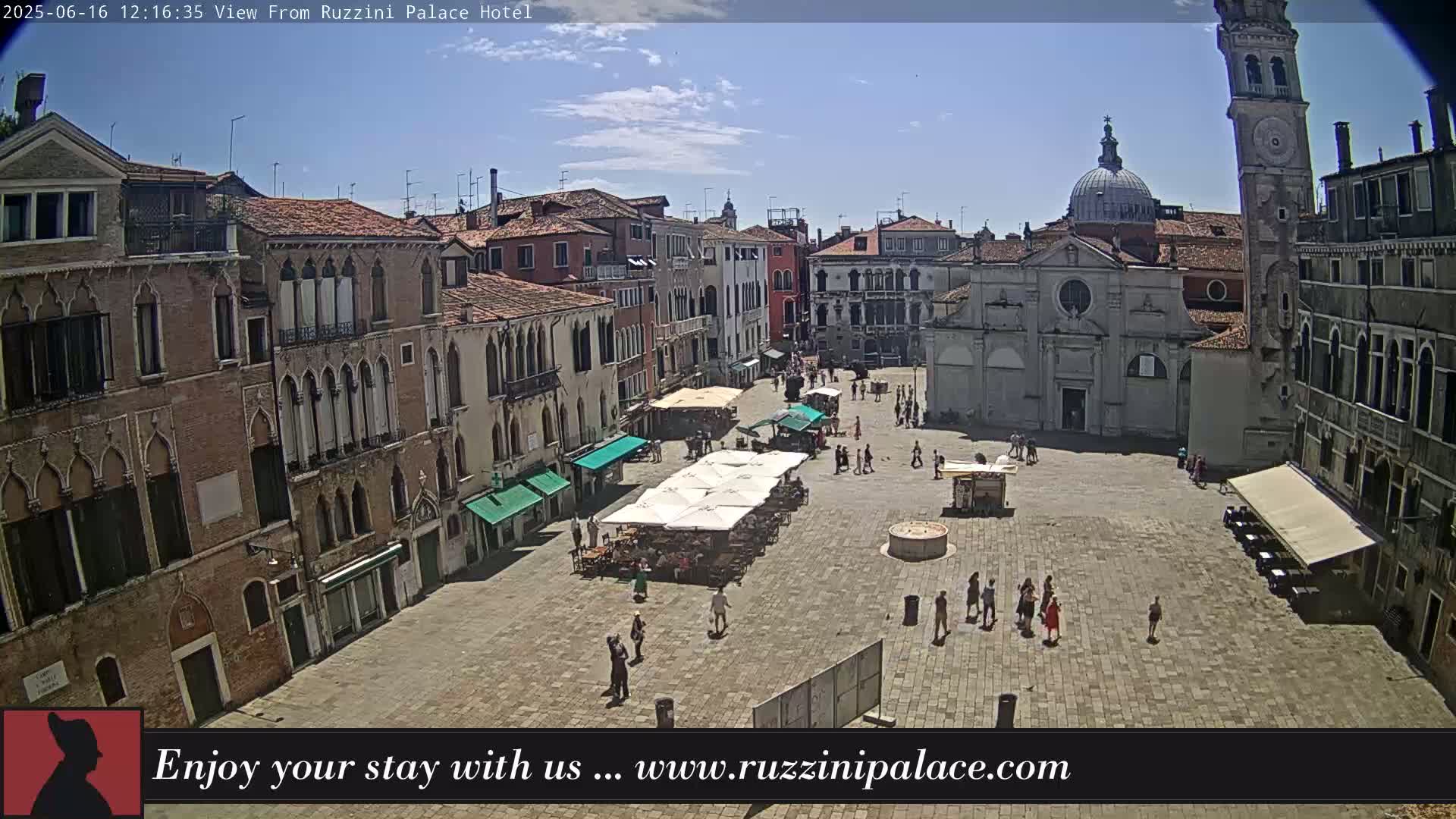 A sunny day shows a large, paved square in Venice, Italy, surrounded by historic buildings, with people strolling and several outdoor cafes set up with umbrellas.