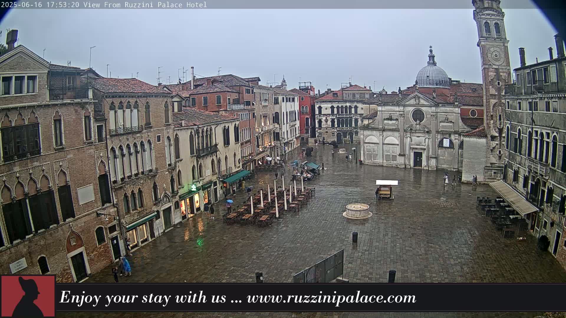 A rain-soaked Venetian piazza is surrounded by old stone buildings, with a few people and empty outdoor seating visible.