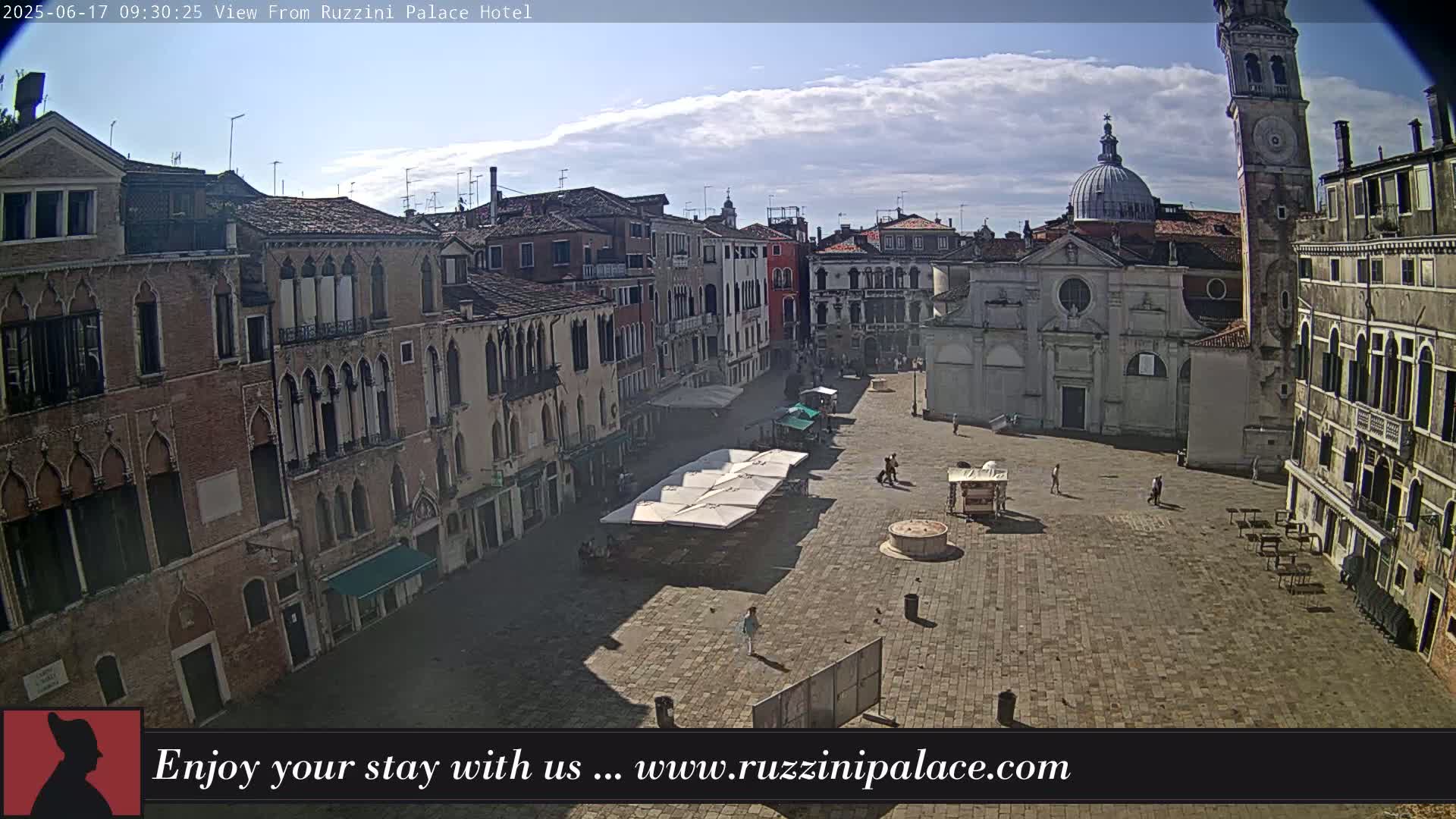 A mostly sunny day reveals a large, paved square in Venice, Italy, surrounded by historic buildings, with a few people and outdoor seating visible.