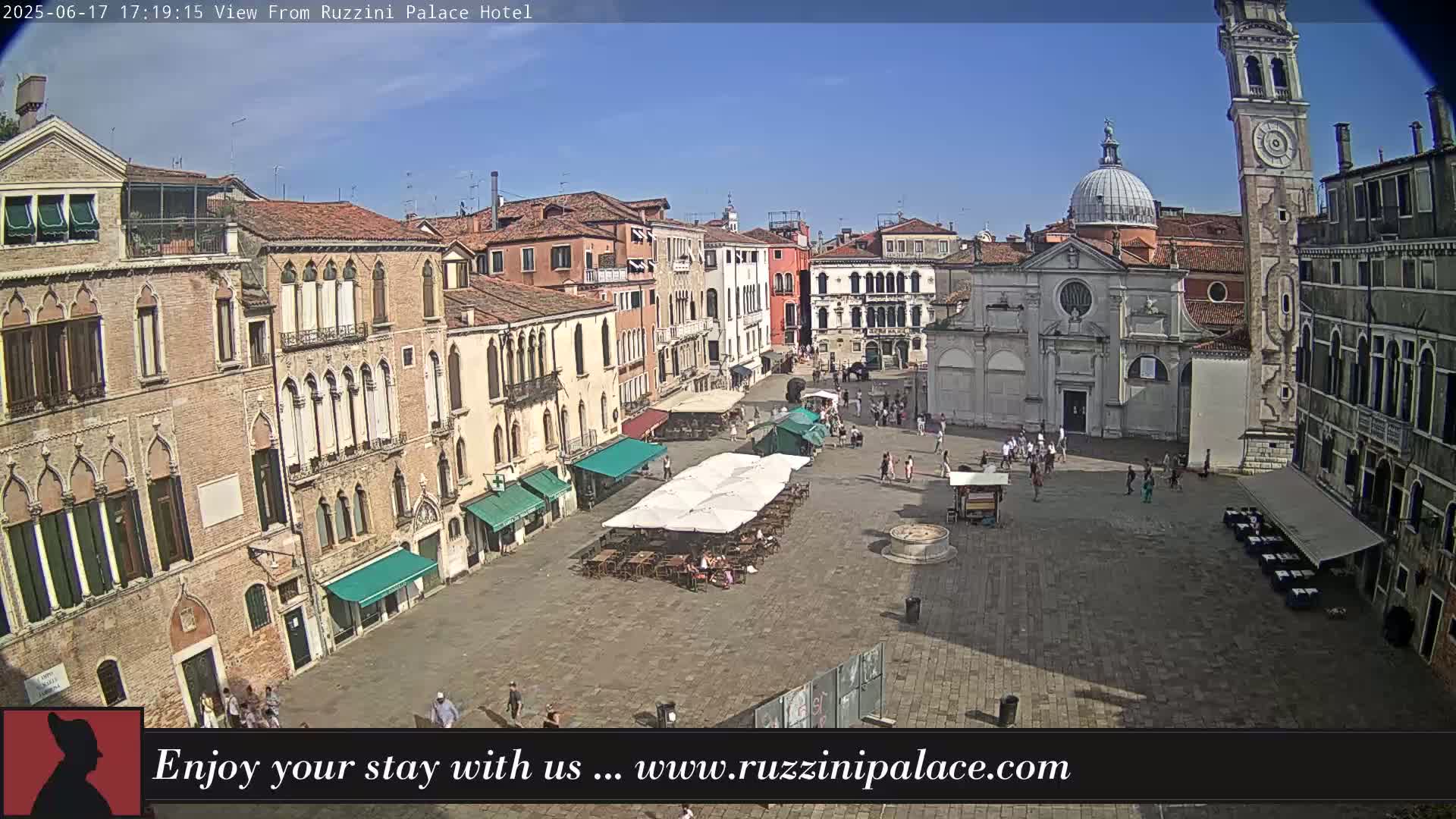 A sunny Venetian piazza is shown, filled with several people, outdoor seating, and old buildings surrounding the square, one of which includes a tall bell tower.