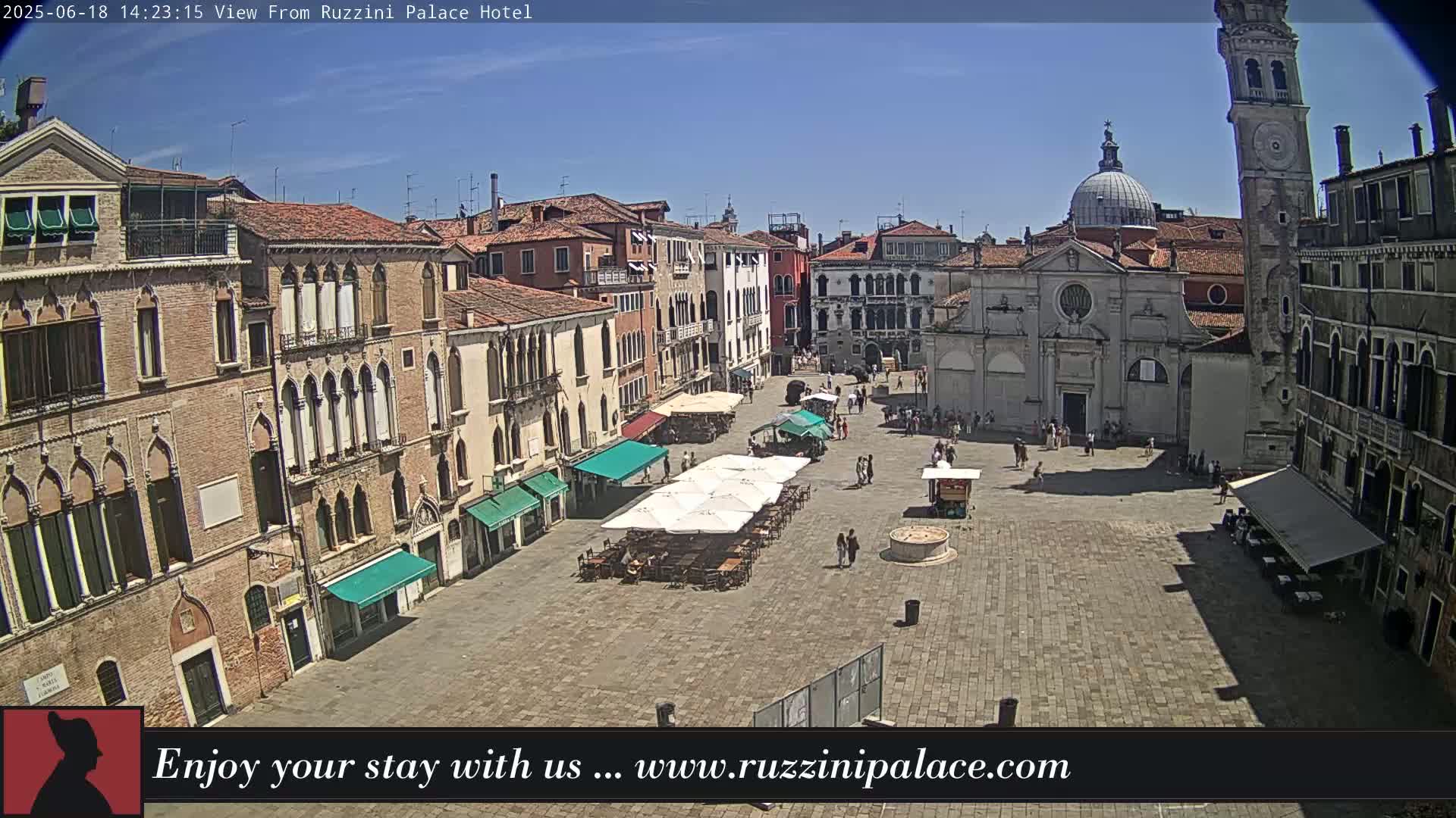 A sunny day in a large, paved square in Venice, Italy, surrounded by historic buildings, with several outdoor cafes set up with umbrellas and tables.