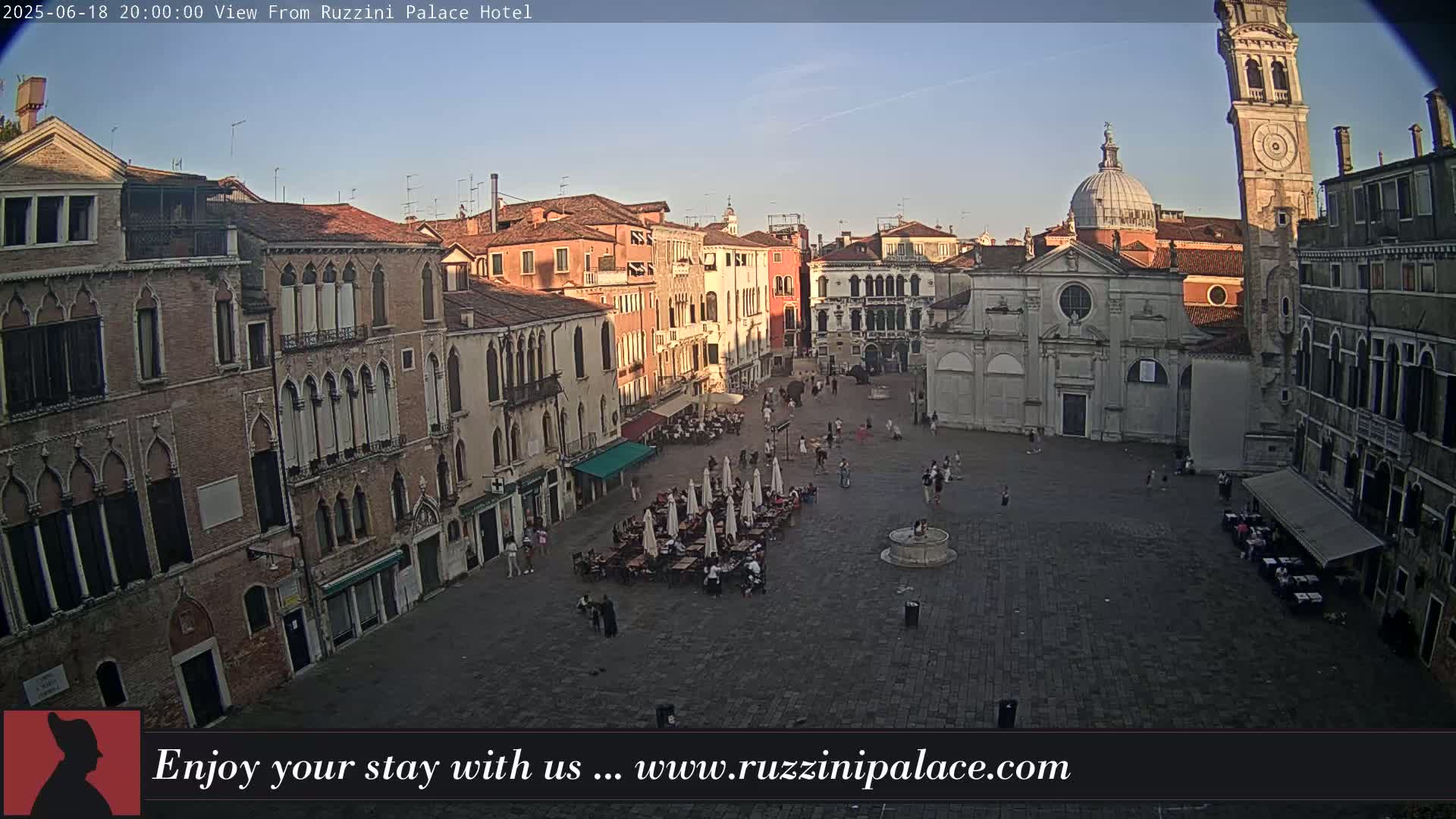 A large, paved square in Venice is filled with several people and outdoor seating under umbrellas, surrounded by historic buildings under a clear evening sky.