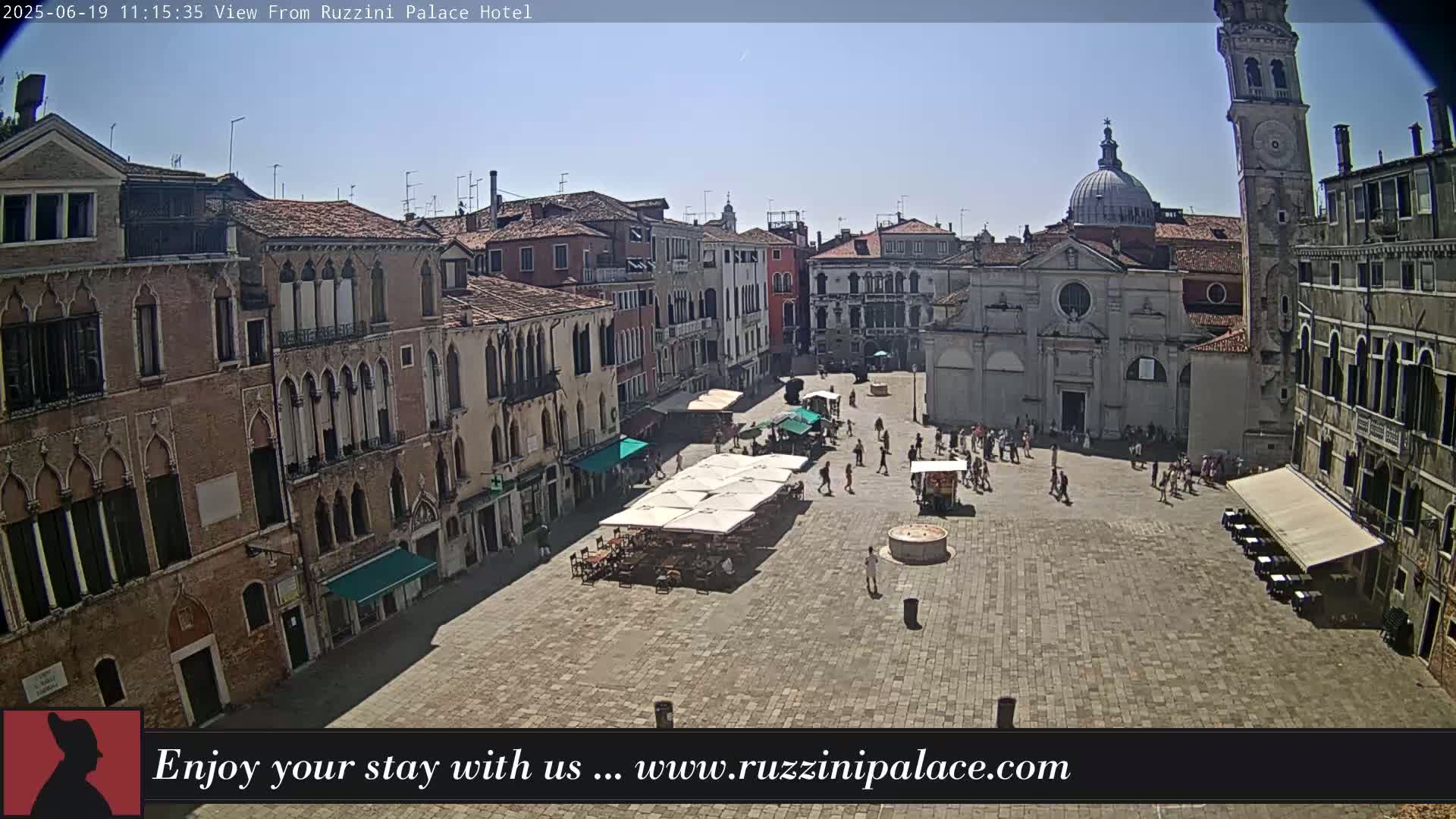 A sunny Venetian square is populated with people and outdoor seating under umbrellas, surrounded by historic buildings, one of which has a tall bell tower.