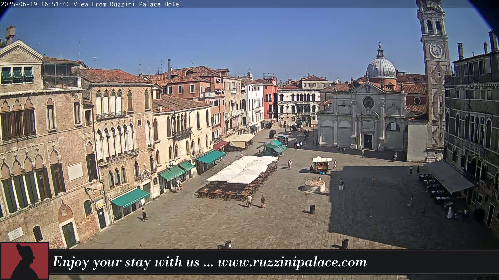 A sunny day in a large, stone plaza in Venice, Italy, with several buildings surrounding it, including a church with a dome and tall bell tower, and outdoor seating under umbrellas.