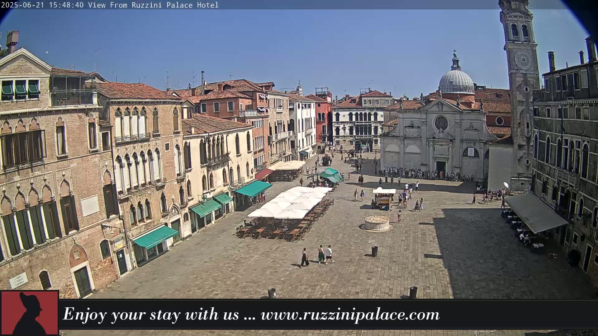 A sunny outdoor view of a large plaza in Venice, Italy, surrounded by historic buildings, with several people and outdoor seating areas.