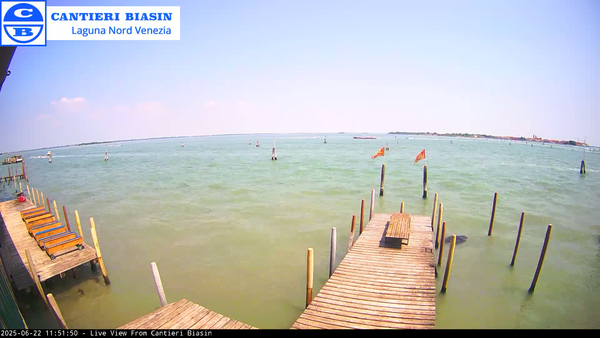 A wooden dock extends over calm, light green water on a sunny day, with a distant landmass visible in the background.
