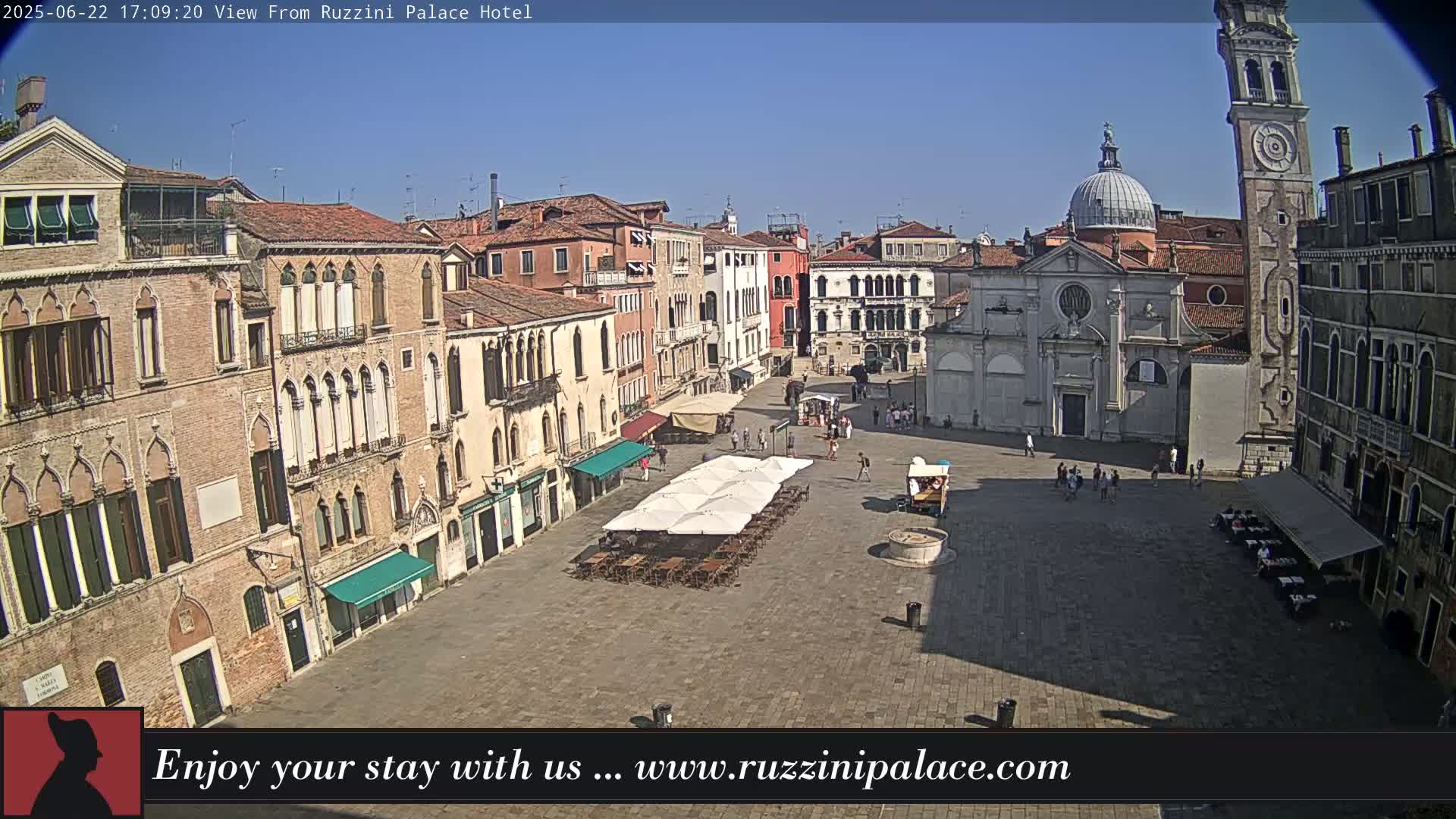 A sunny day in a large, paved square in Venice, Italy, surrounded by old buildings, with several people and outdoor seating under umbrellas.