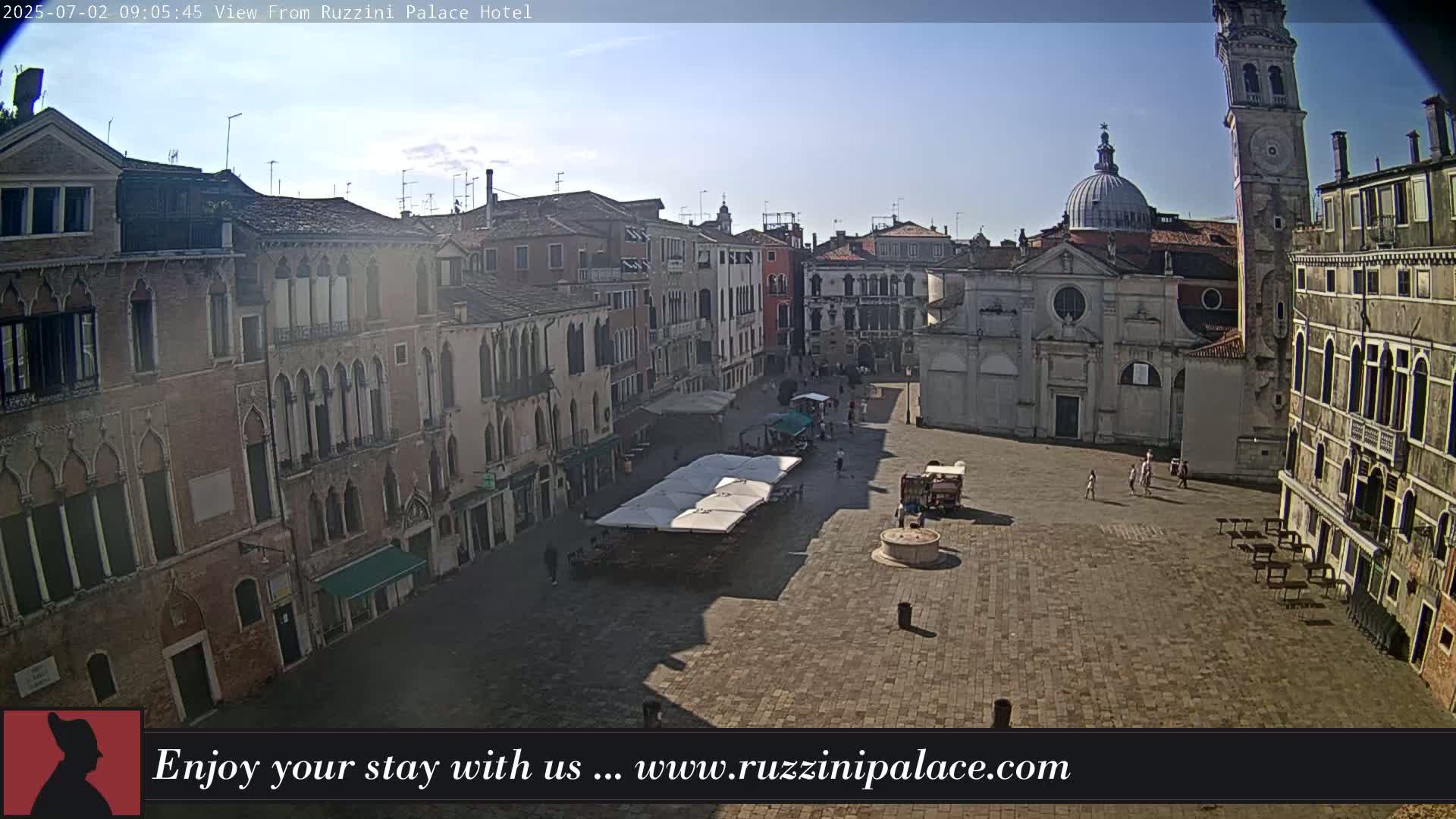 A sunny day reveals a large, mostly empty plaza in Venice, surrounded by historic buildings, including a church with a dome and a tall bell tower.