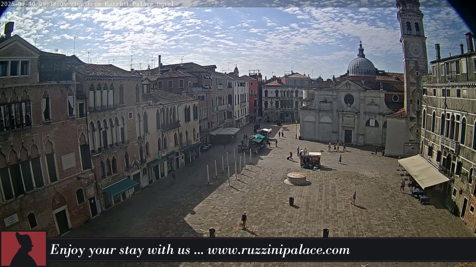 A sunny day reveals a large, mostly empty cobblestone square in Venice, surrounded by ancient buildings, including a church with a dome and a tall clock tower.