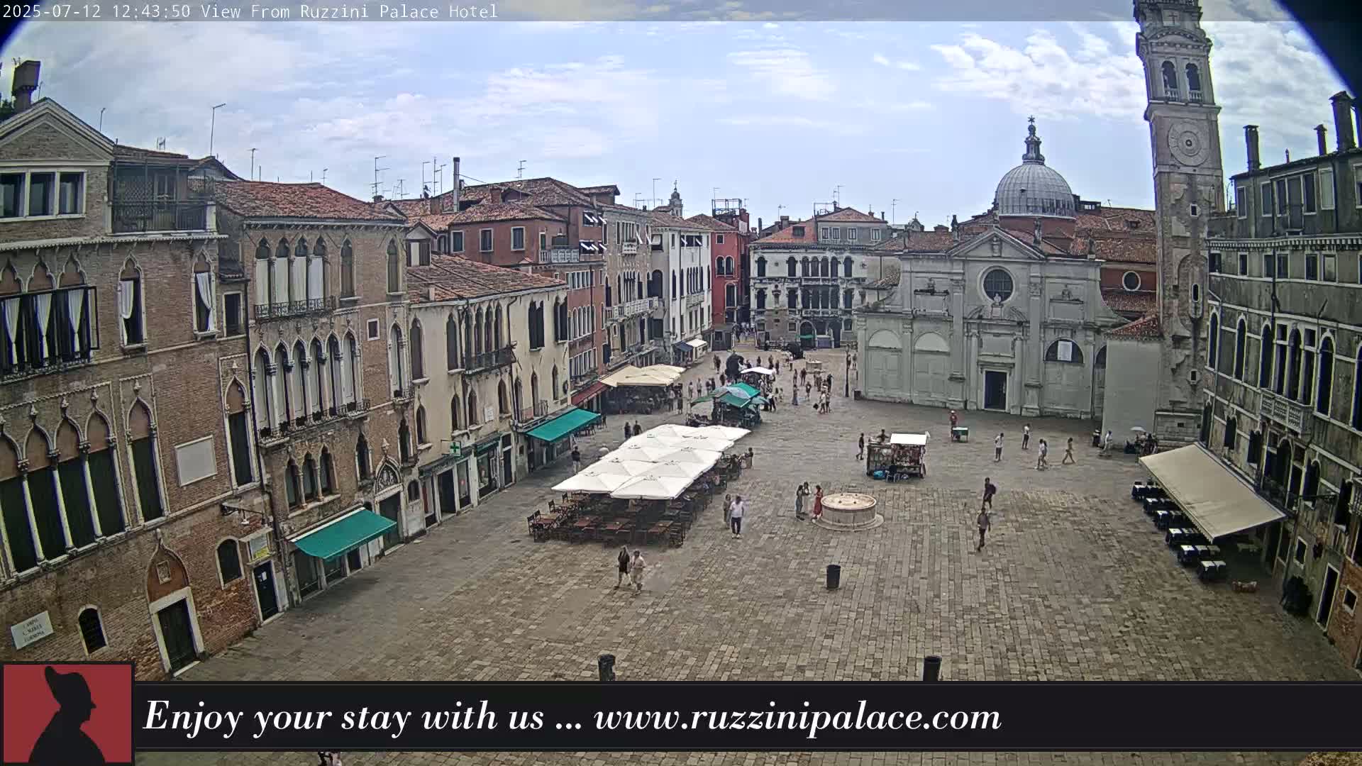 A sunny day reveals a large, mostly empty cobblestone square in Venice, surrounded by ancient buildings, including a church with a dome and a tall clock tower.