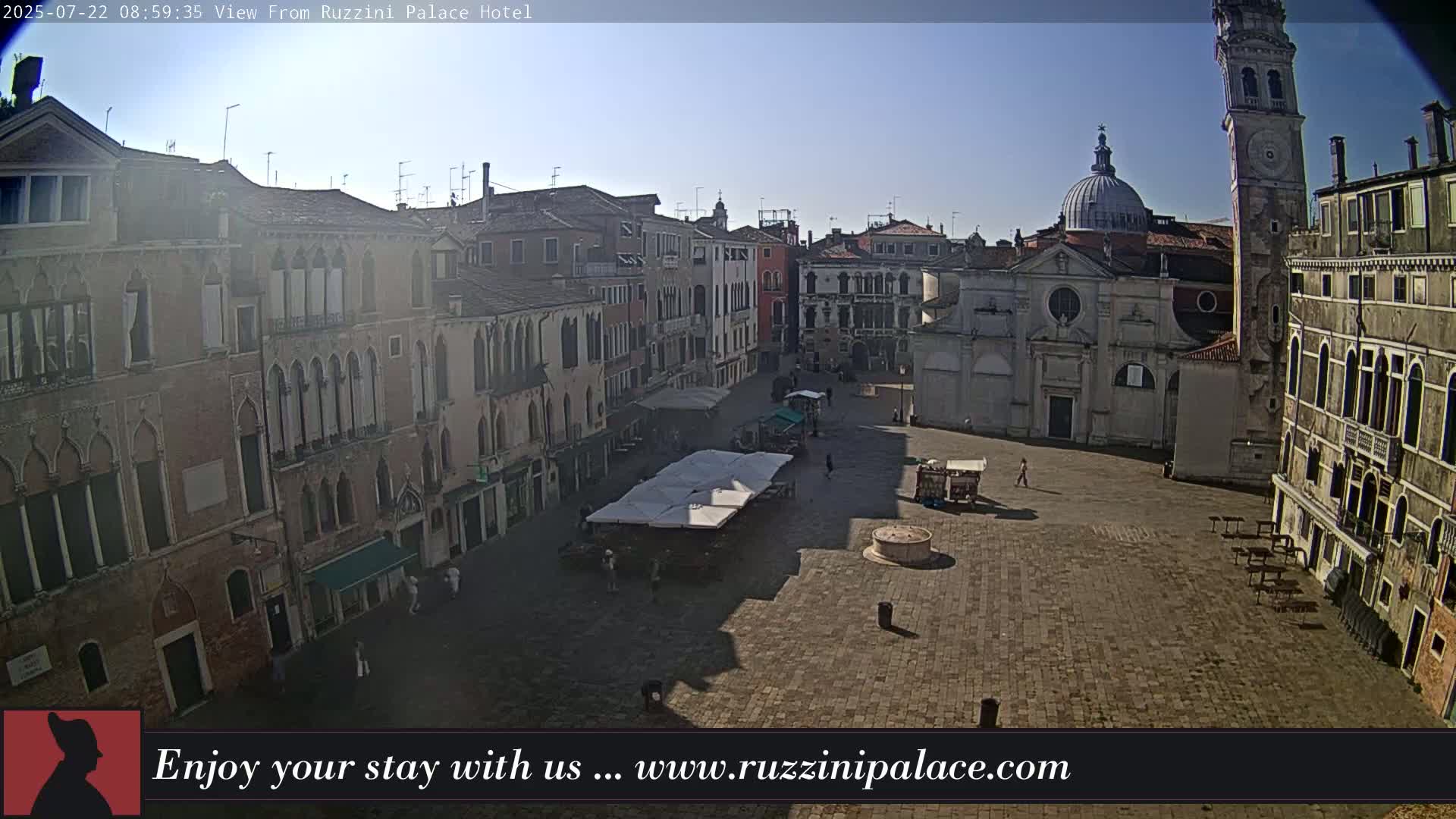 A sunny view of a large, mostly empty square in Venice, Italy, surrounded by historic buildings, including a church and a tall tower.