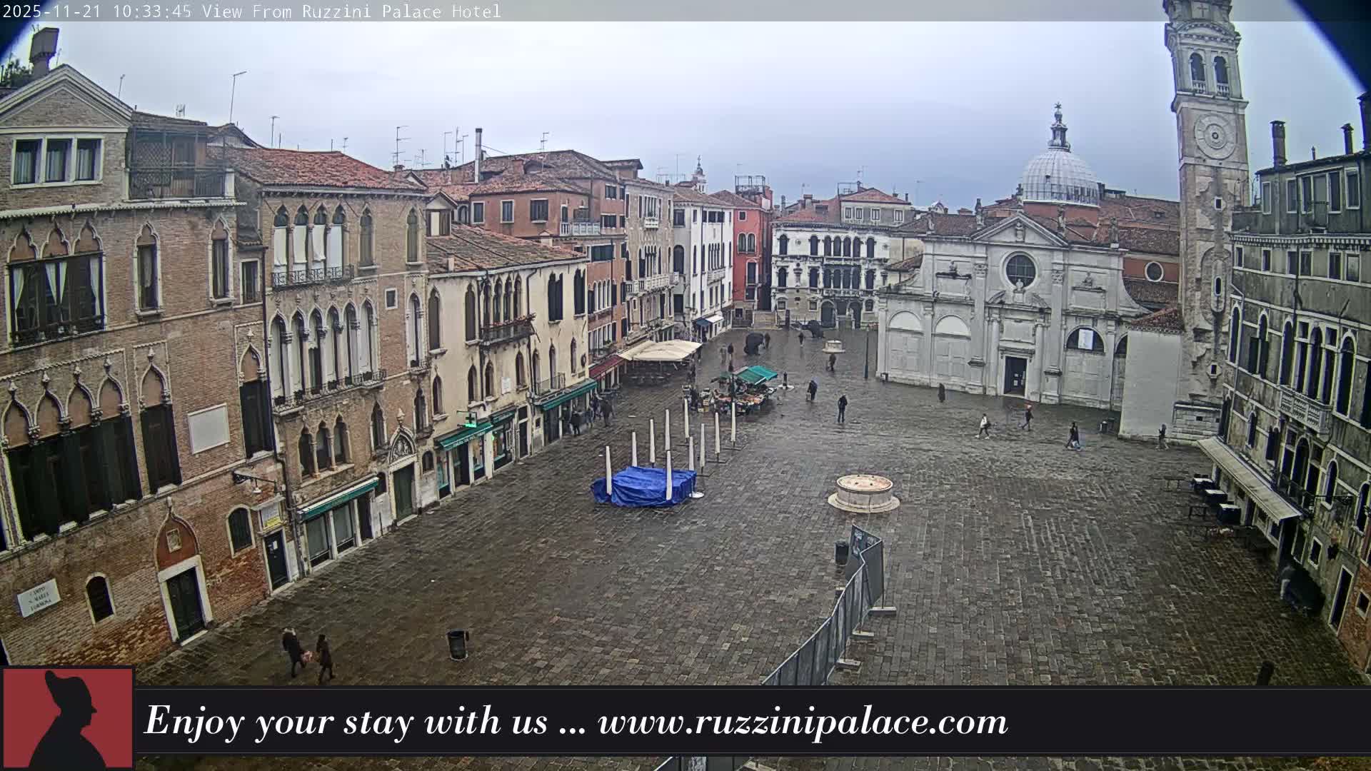 A wide aerial view captures a historic European piazza, featuring a prominent church with a dome and an adjacent bell tower, surrounded by traditional buildings, as several people traverse the wet, cobblestone ground under an overcast sky.