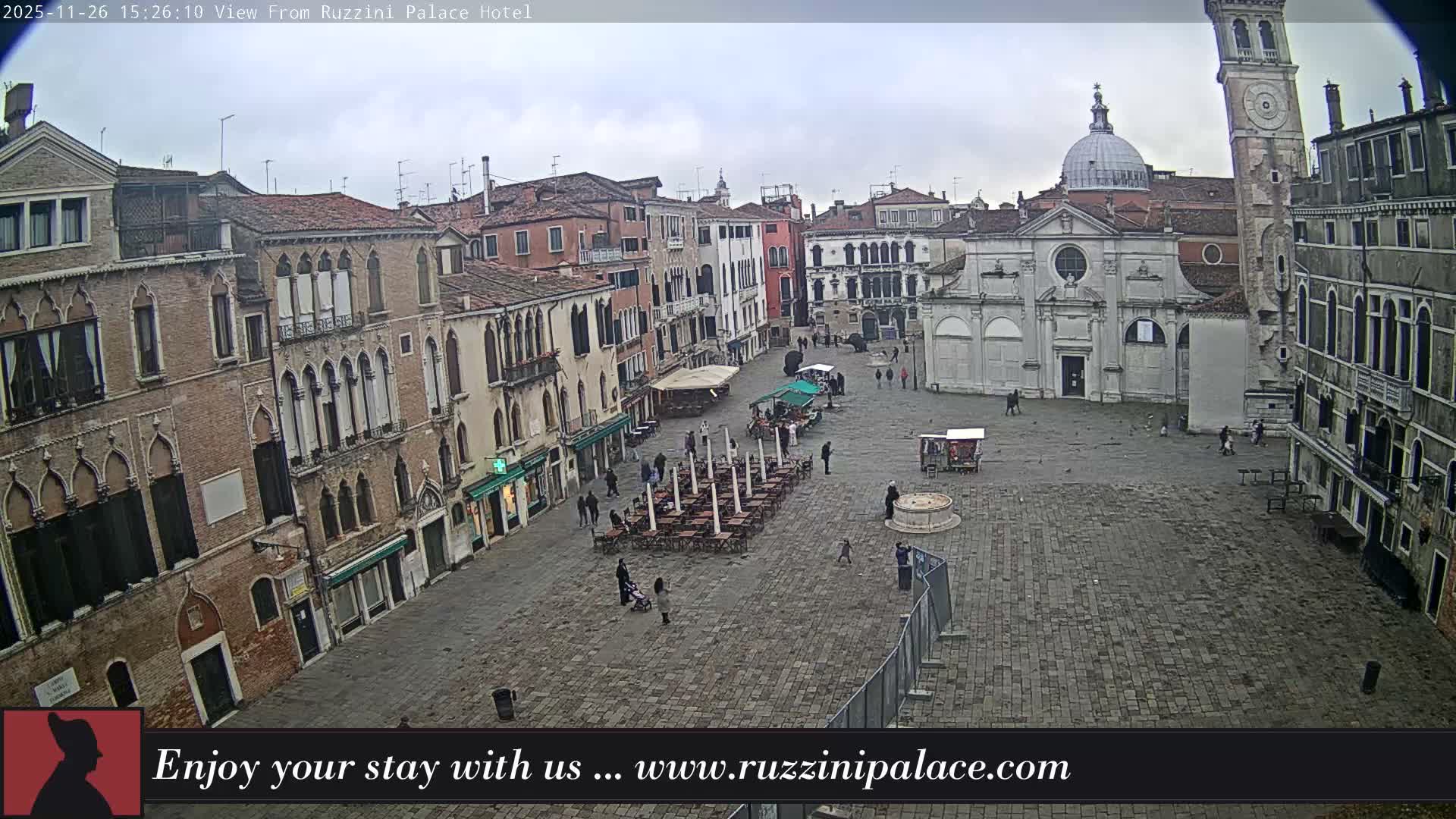 A wide aerial view captures a historic European piazza, featuring a prominent church with a dome and an adjacent bell tower, surrounded by traditional buildings, as several people traverse the wet, cobblestone ground under an overcast sky.
