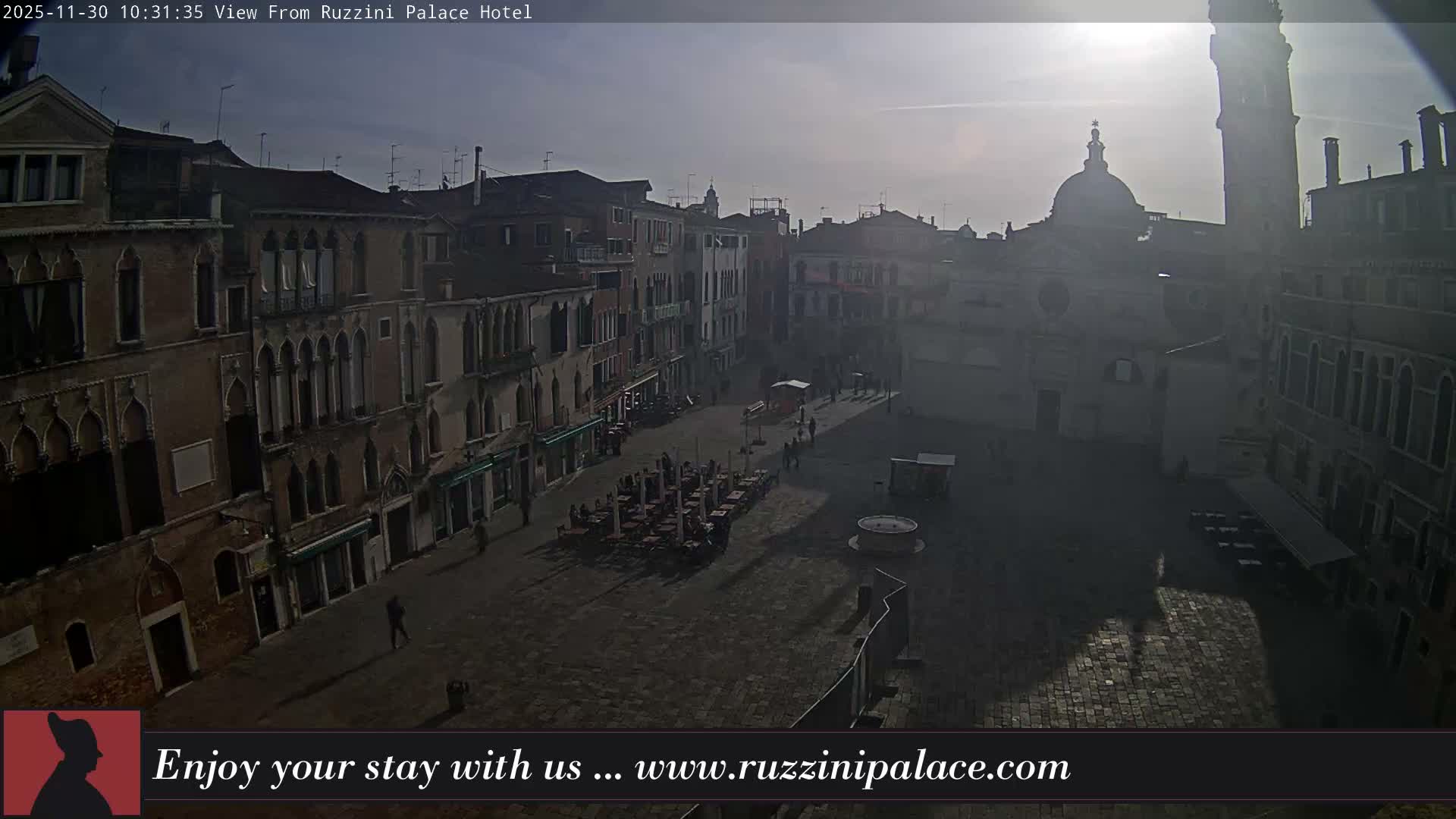 An aerial view captures a historic Venetian piazza lined with ornate multi-story buildings and an active outdoor cafe, with a prominent church dome and tall bell tower in the background, all bathed in bright, hazy sunlight casting long shadows.