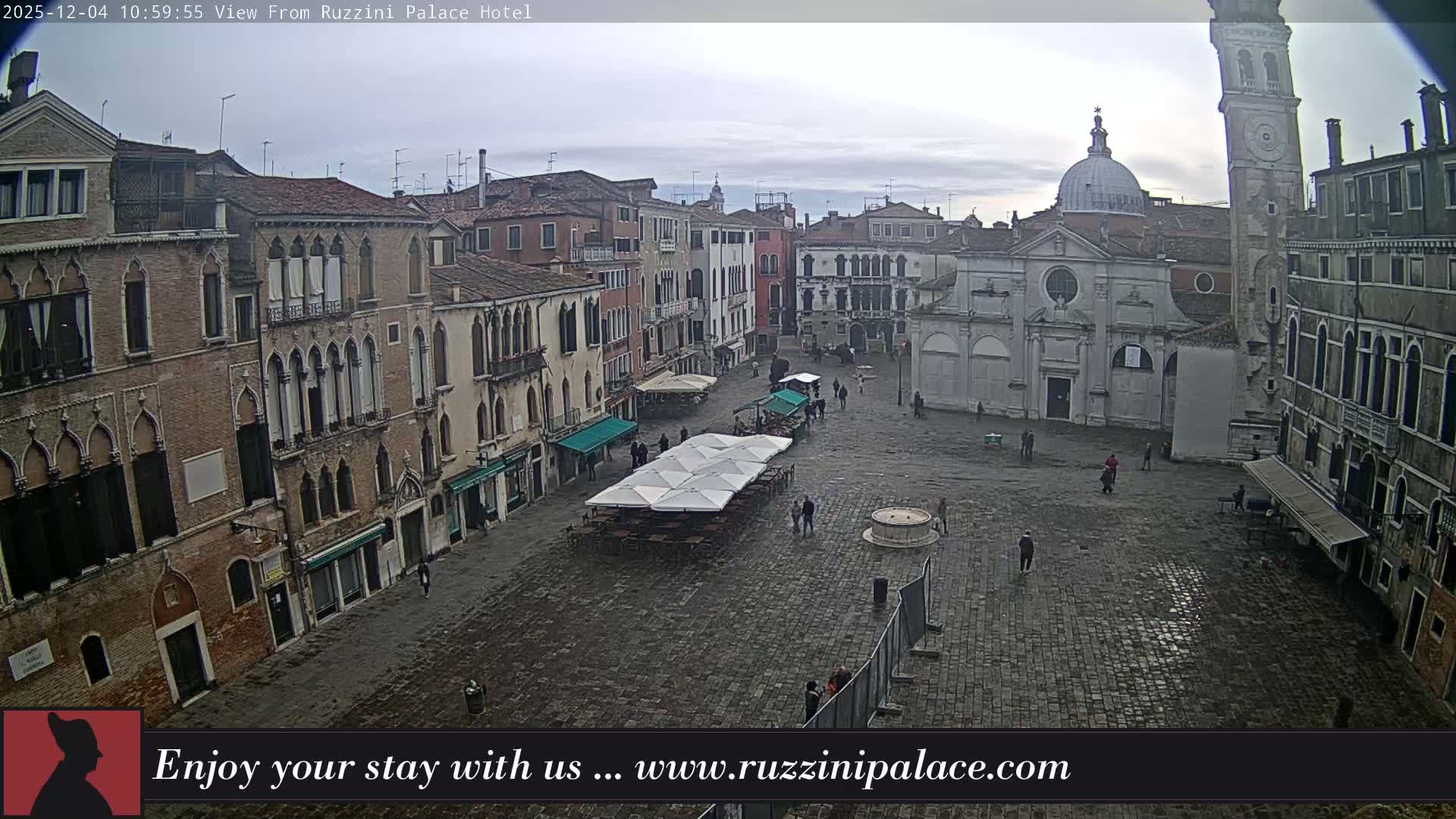 A wide-angle view reveals a historic European piazza, featuring a prominent domed church and a tall bell tower, surrounded by multi-story buildings and outdoor cafe seating, with a few pedestrians on the damp cobblestone ground under an overcast sky.