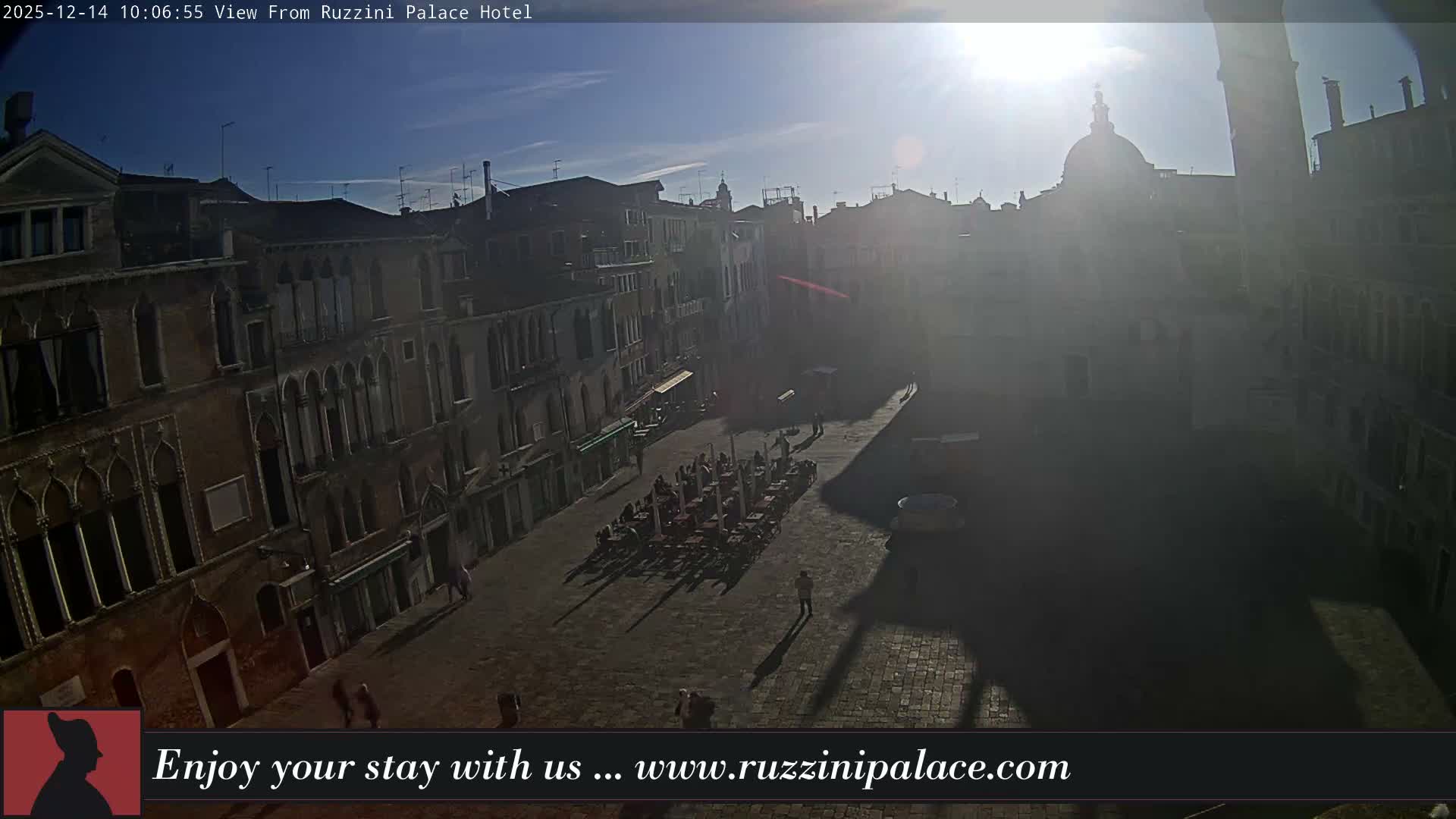 A wide-angle view reveals a historic European piazza, featuring a prominent domed church and a tall bell tower, surrounded by multi-story buildings and outdoor cafe seating, with a few pedestrians on the damp cobblestone ground under an overcast sky.