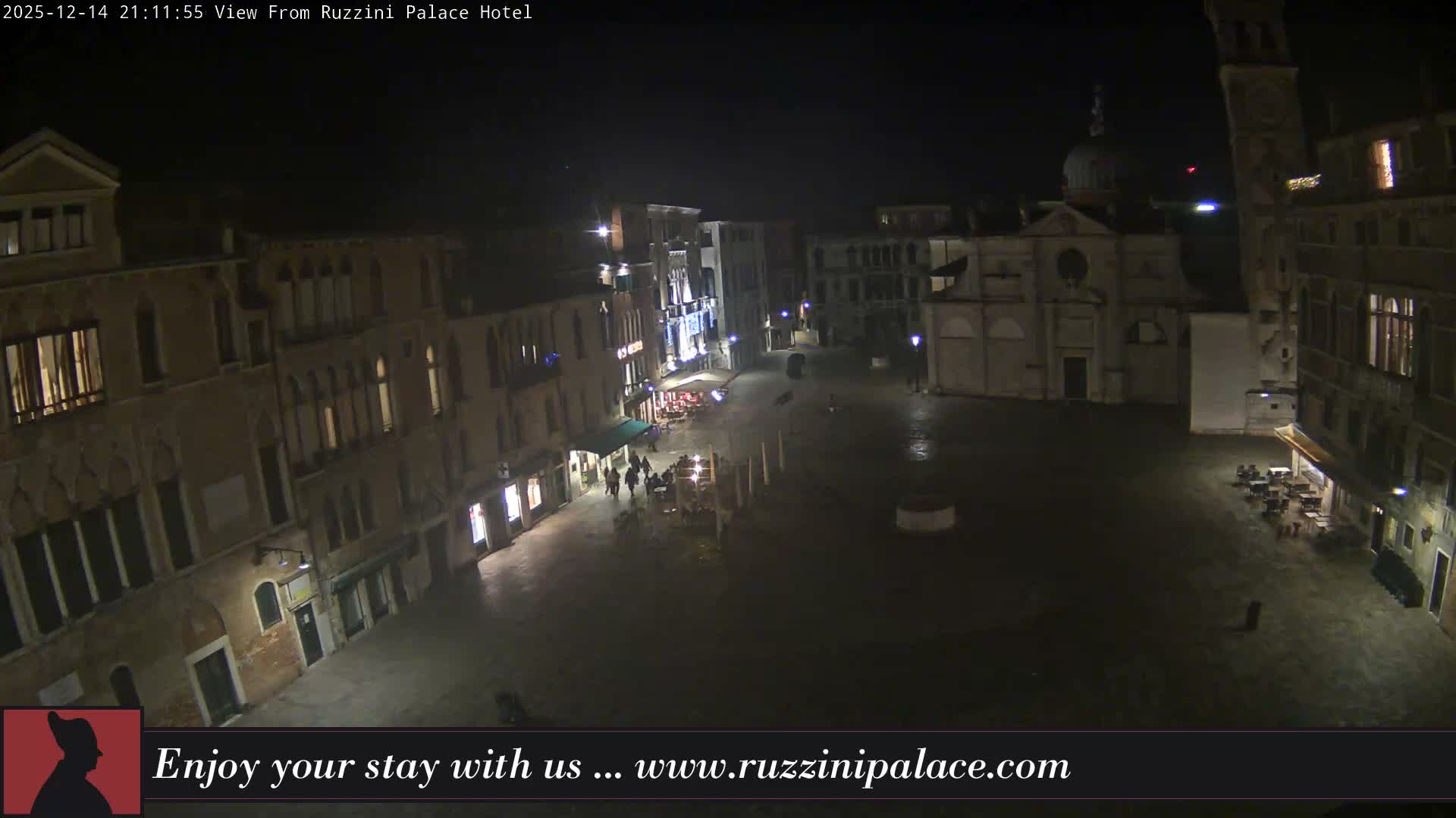 A wide-angle view reveals a historic European piazza, featuring a prominent domed church and a tall bell tower, surrounded by multi-story buildings and outdoor cafe seating, with a few pedestrians on the damp cobblestone ground under an overcast sky.